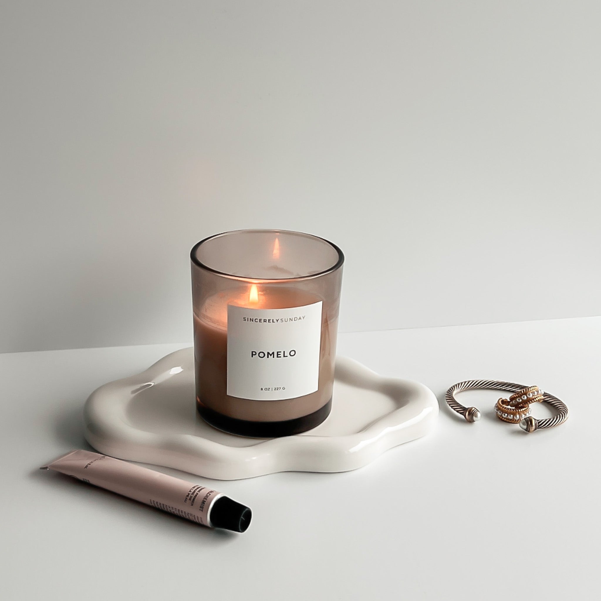 Pomelo candle on a white cloud-shaped tray, styled with a tube of lip gloss and jewelry in a decorative setup.