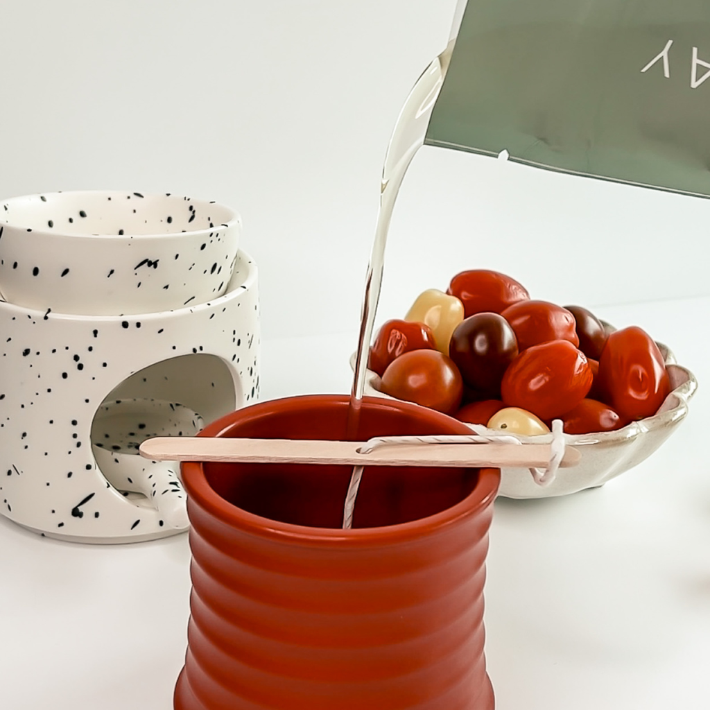 Wax refill being poured into a red vessel, with a wax warmer and a bowl of fresh tomatoes in the background.
