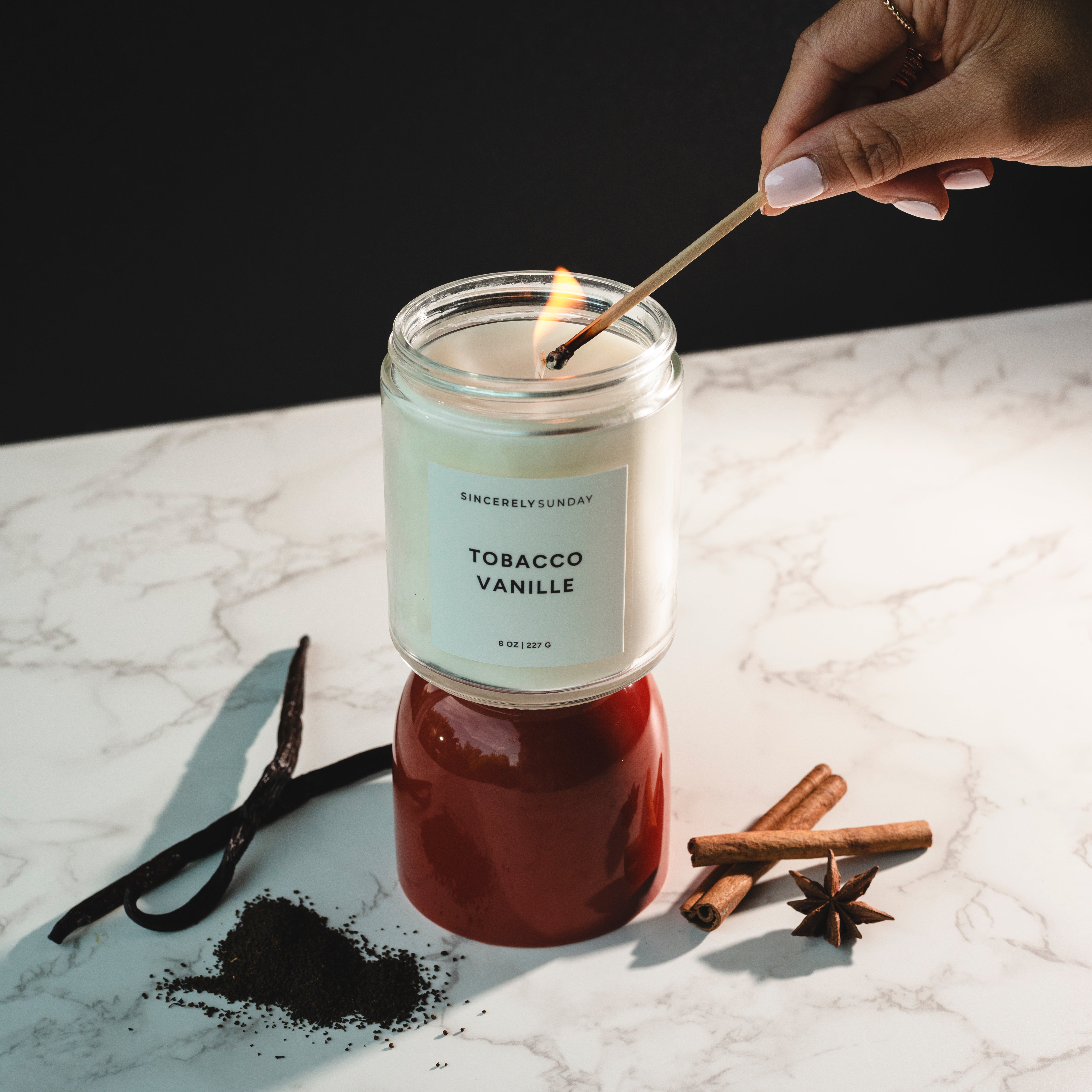 Candle labeled 'Tobacco Vanille' being lit on a marble surface with vanilla bean, cinnamon stick, and star anise.