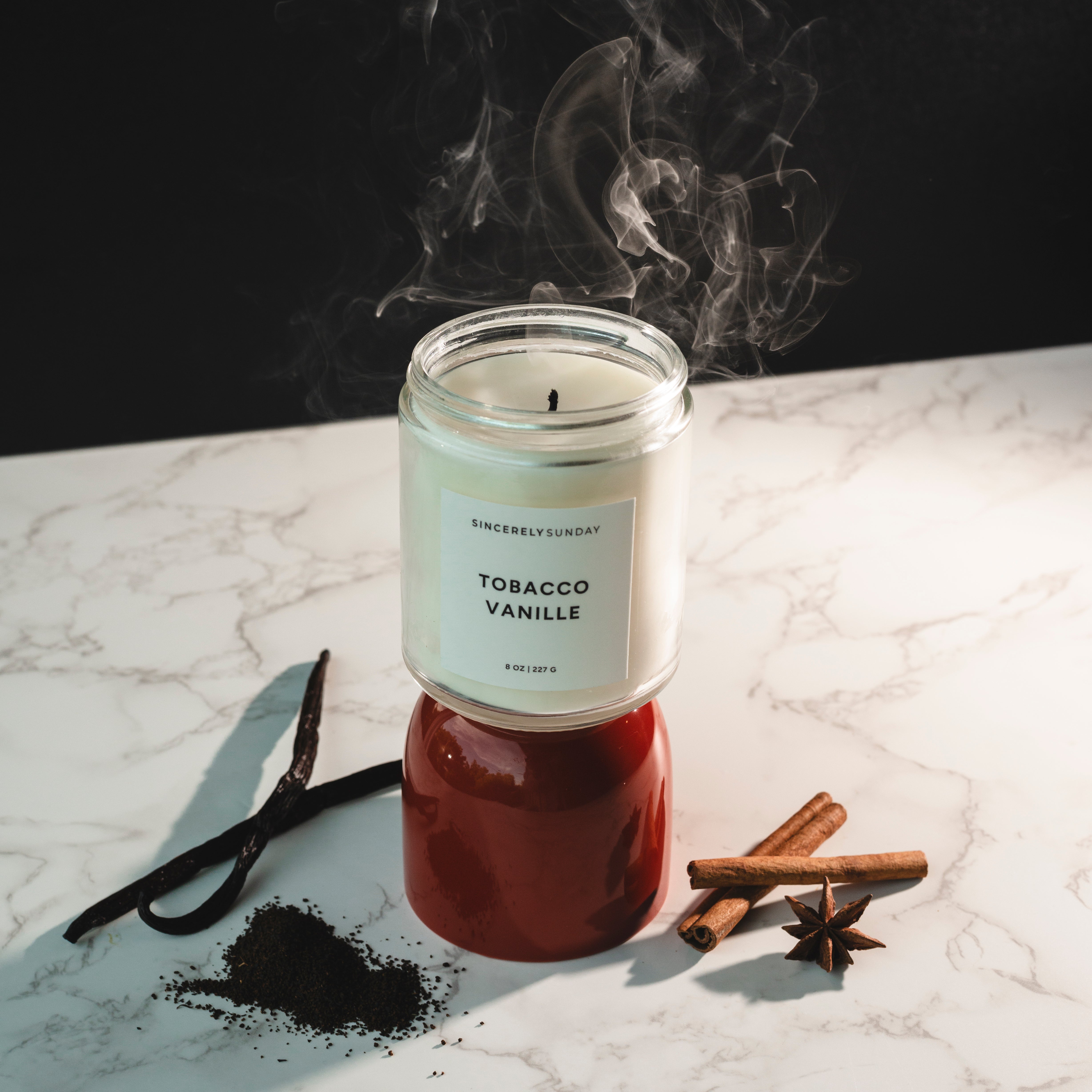 Tobacco Vanille candle on a red base with smoke rising, placed beside spices on a dark background.