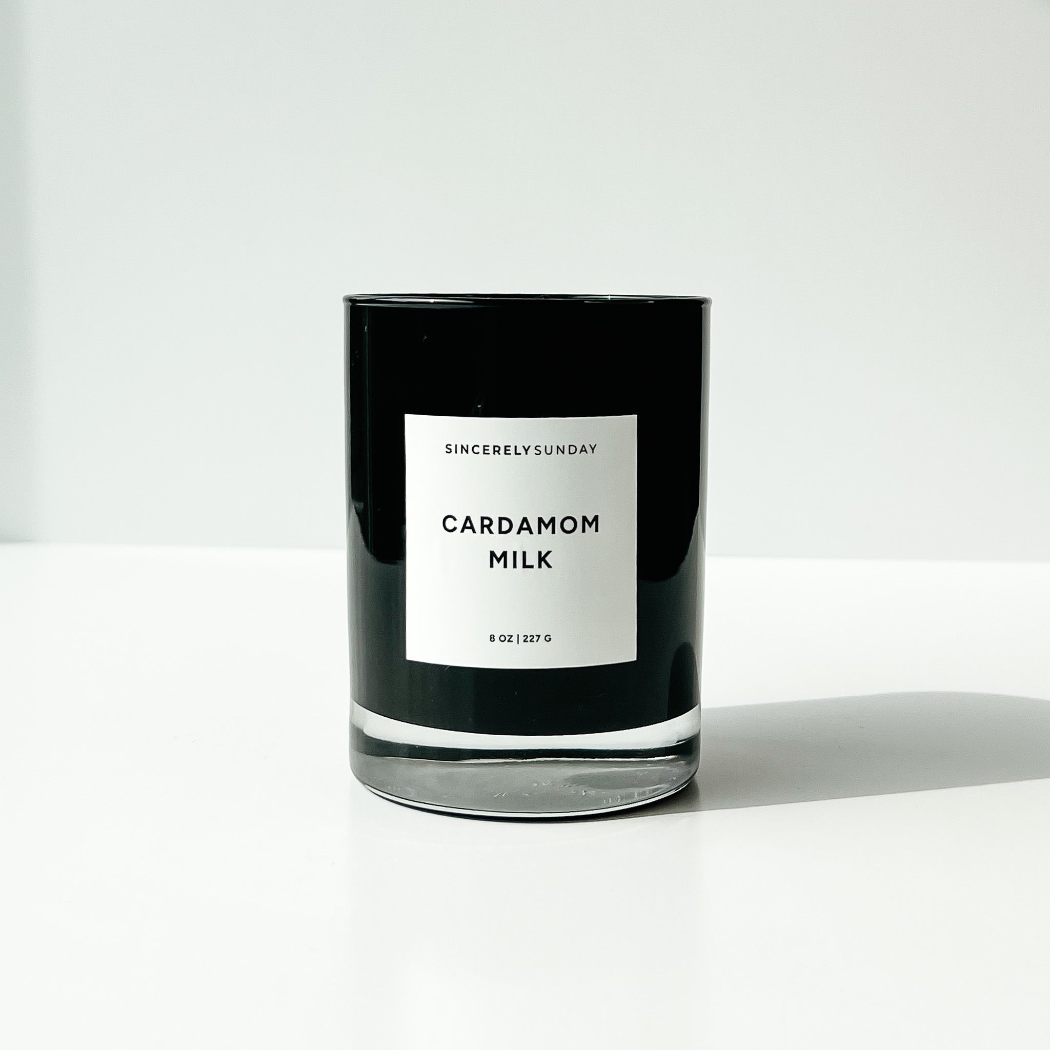 Black candle labeled 'Cardamom Milk' by 'Sincerely Sunday' on a white background