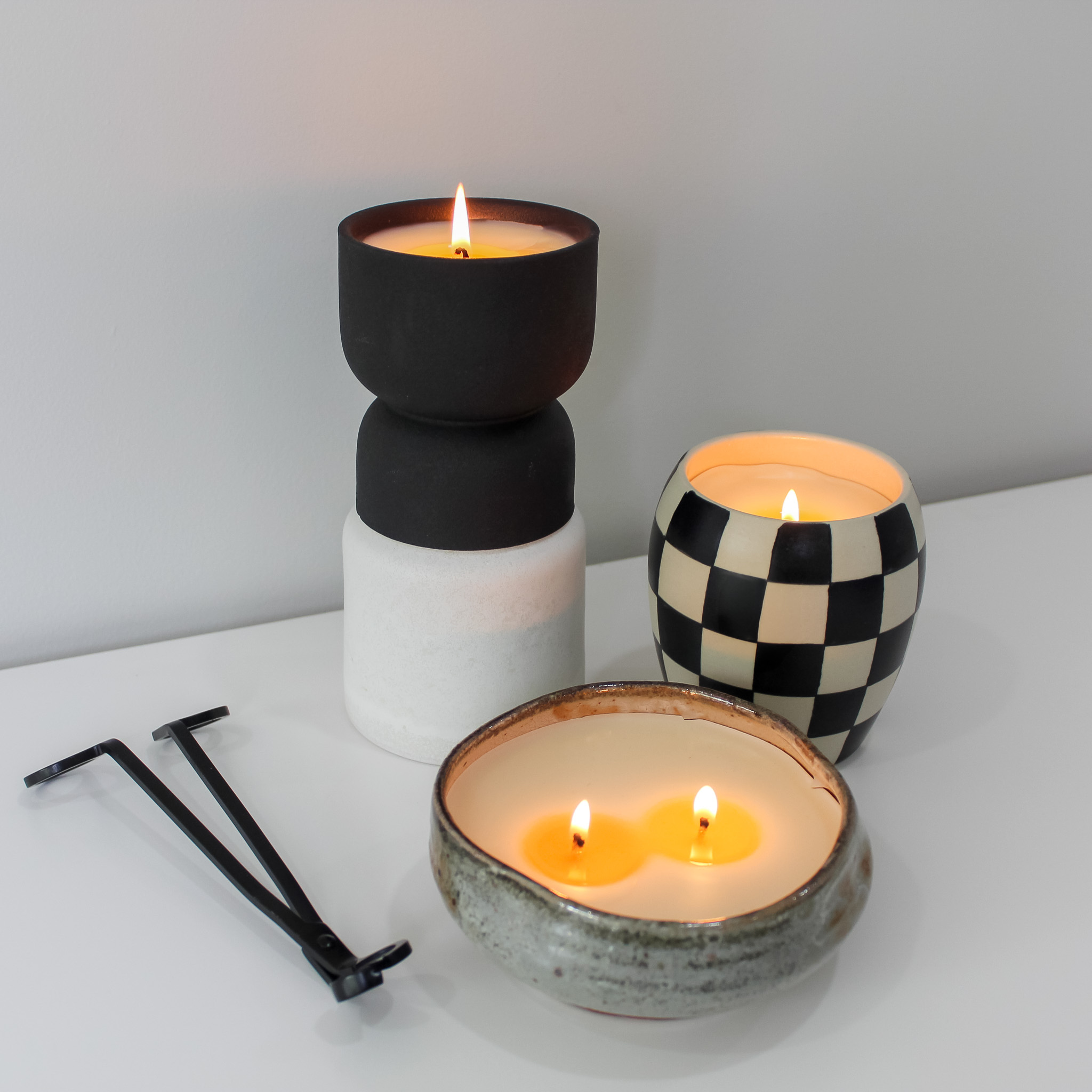 Three lit black and white decorative candles beside a black wick trimmer on a white surface.