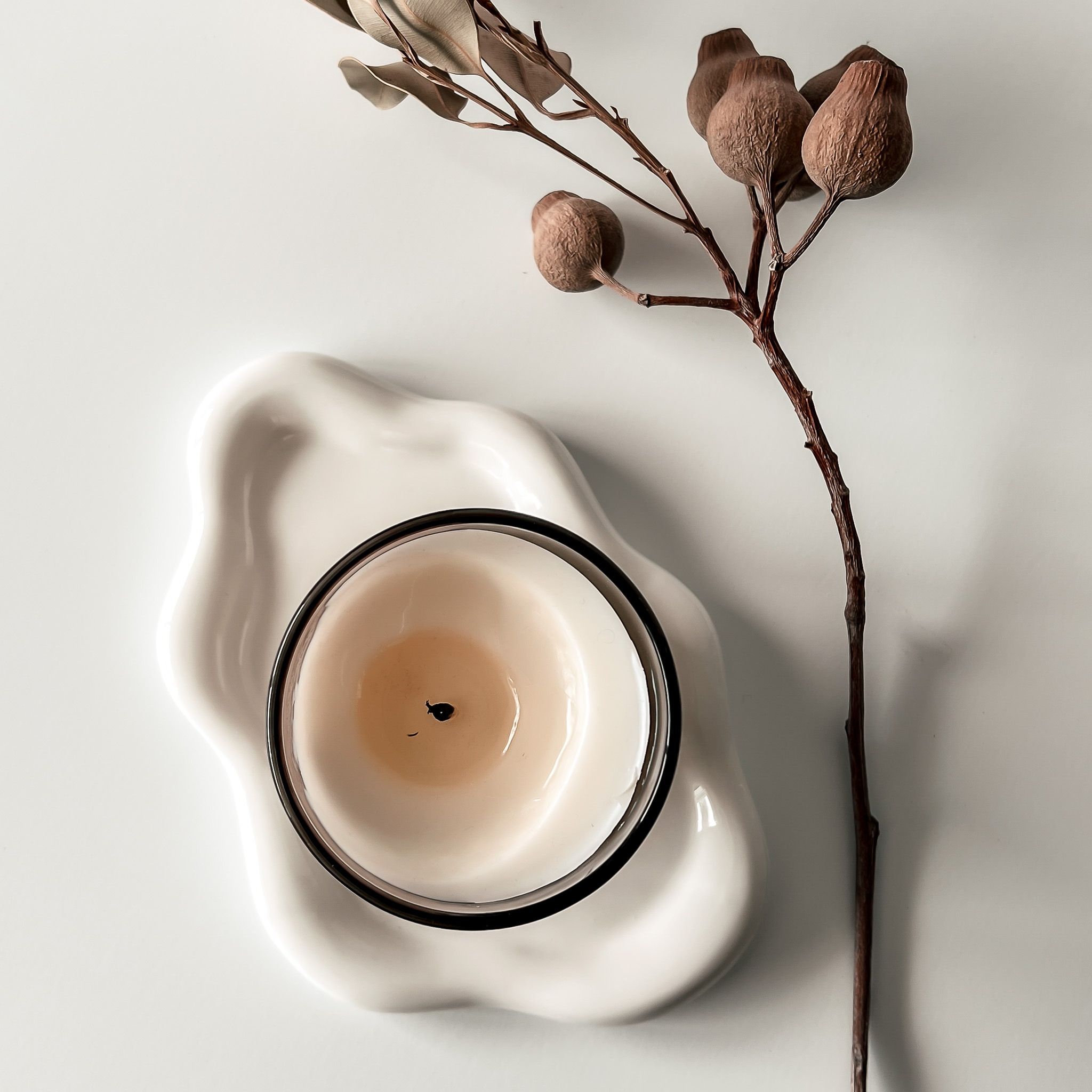 Top-down view of a candle with excess wax on the sides, placed on a white scalloped dish beside an olive branch.