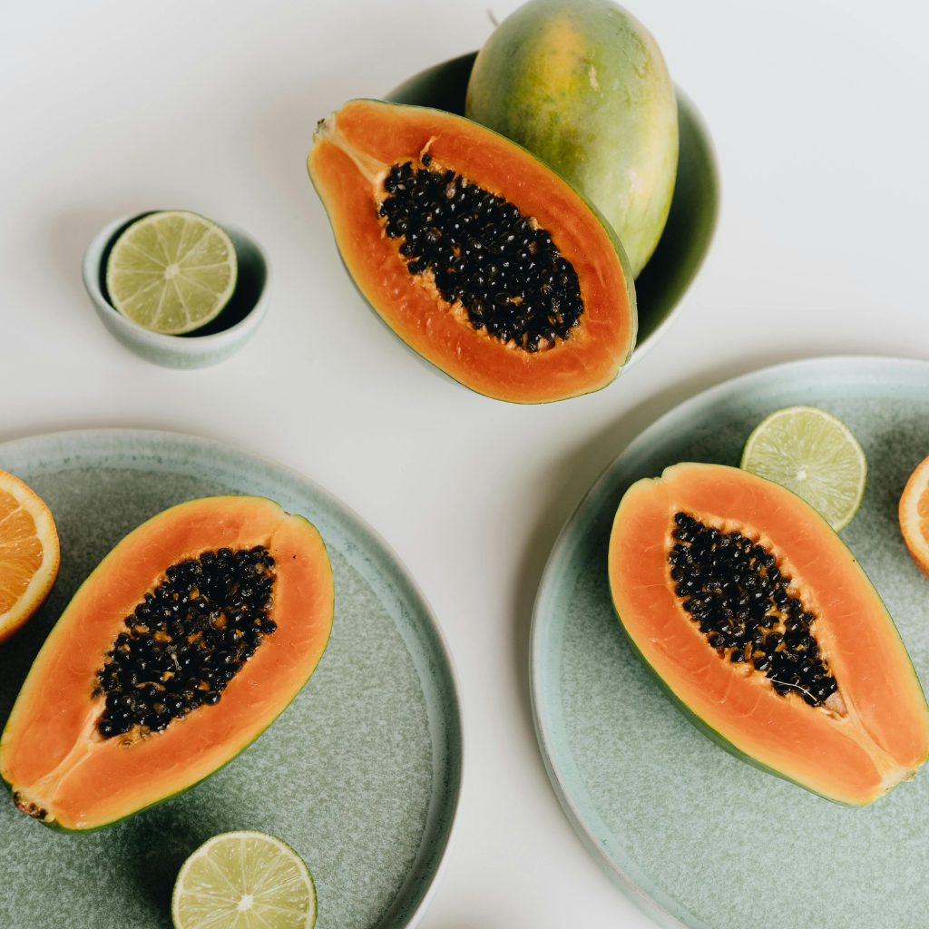 A beautifully arranged overhead shot of papaya and limes on speckled sage green plates, some whole, some sliced open, radiating lush, juicy freshness.