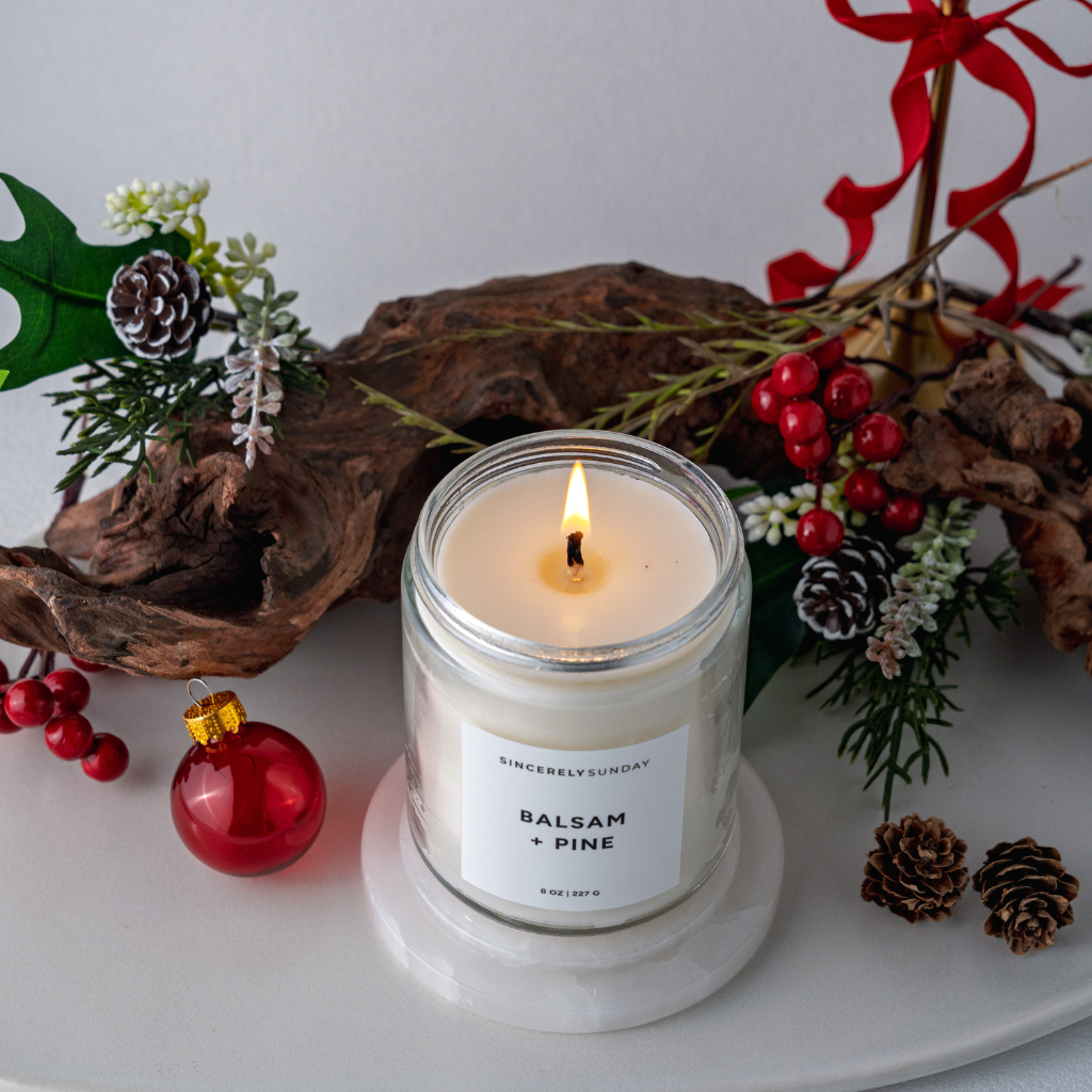 Candle labeled 'Balsam + Pine' surrounded by holiday decorations on a white surface