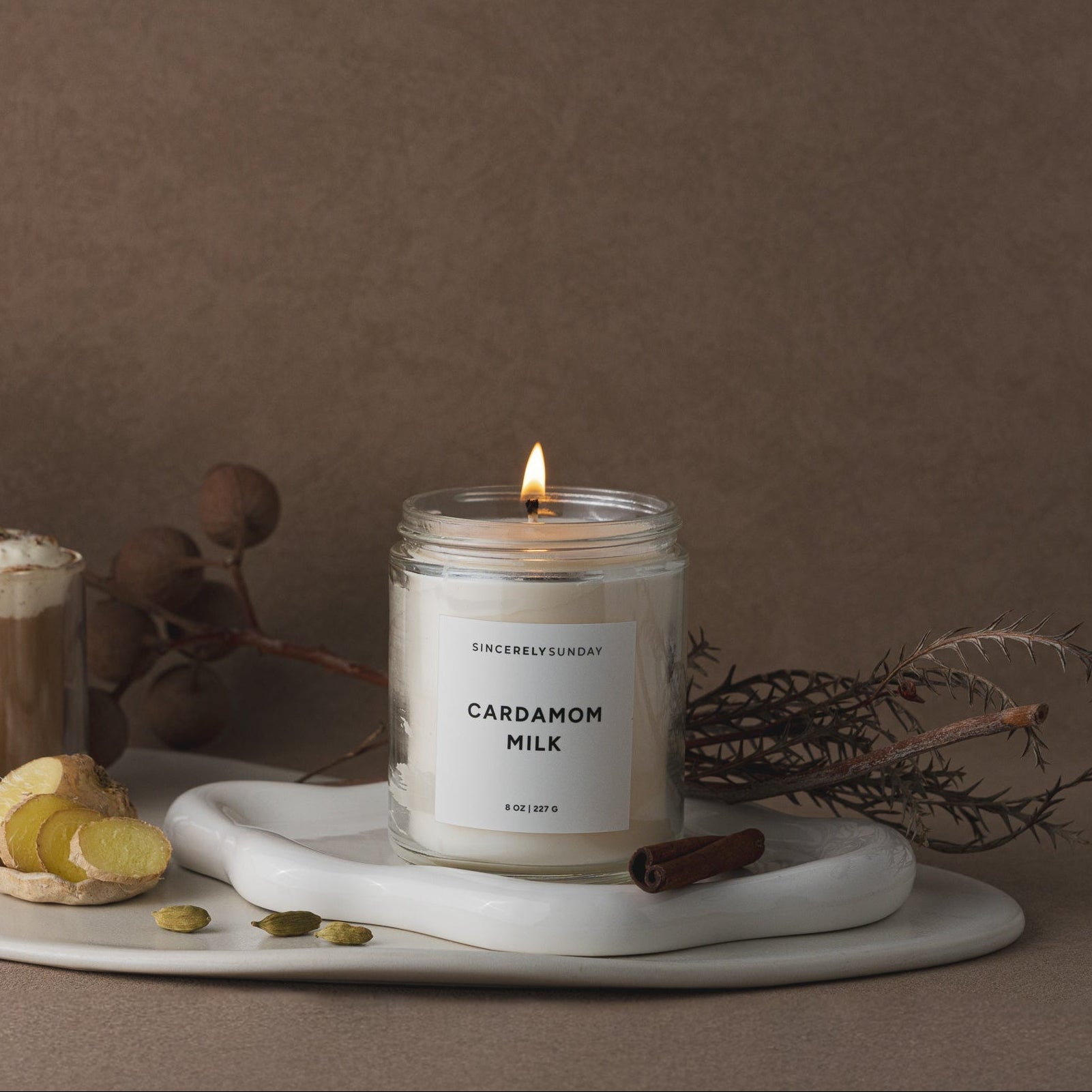 Candle labeled 'Cardamom Milk' on a marble tray with a drink and garnishes against a brown background