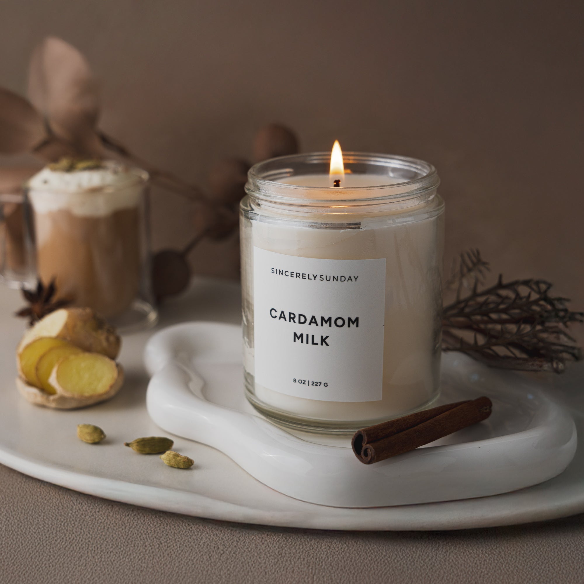 Candle labeled 'Cardamom Milk' on a tray with a glass of hot chocolate and garnishes.