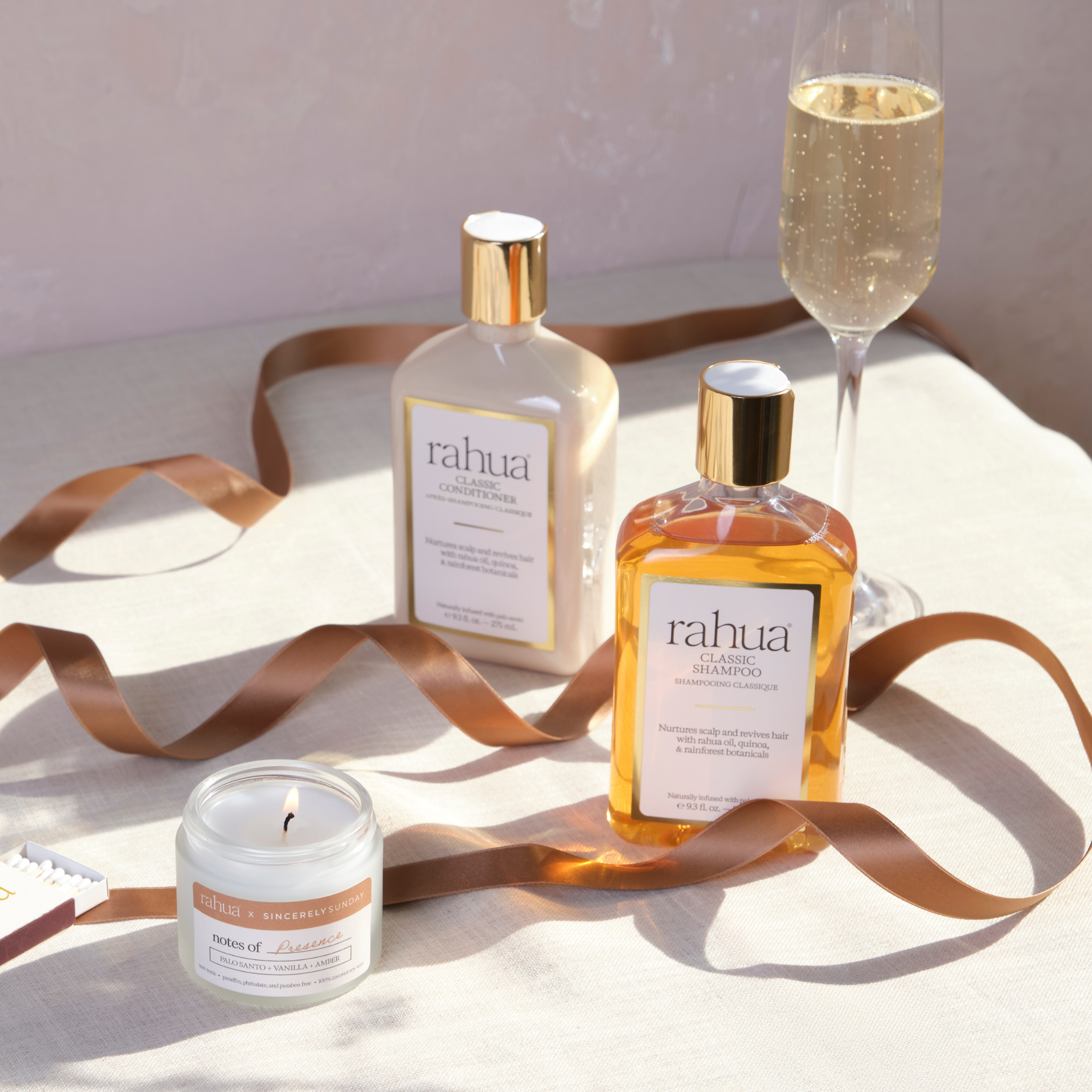 Two bottles of rahua hair care products with a candle and glass of champagne on a light surface.