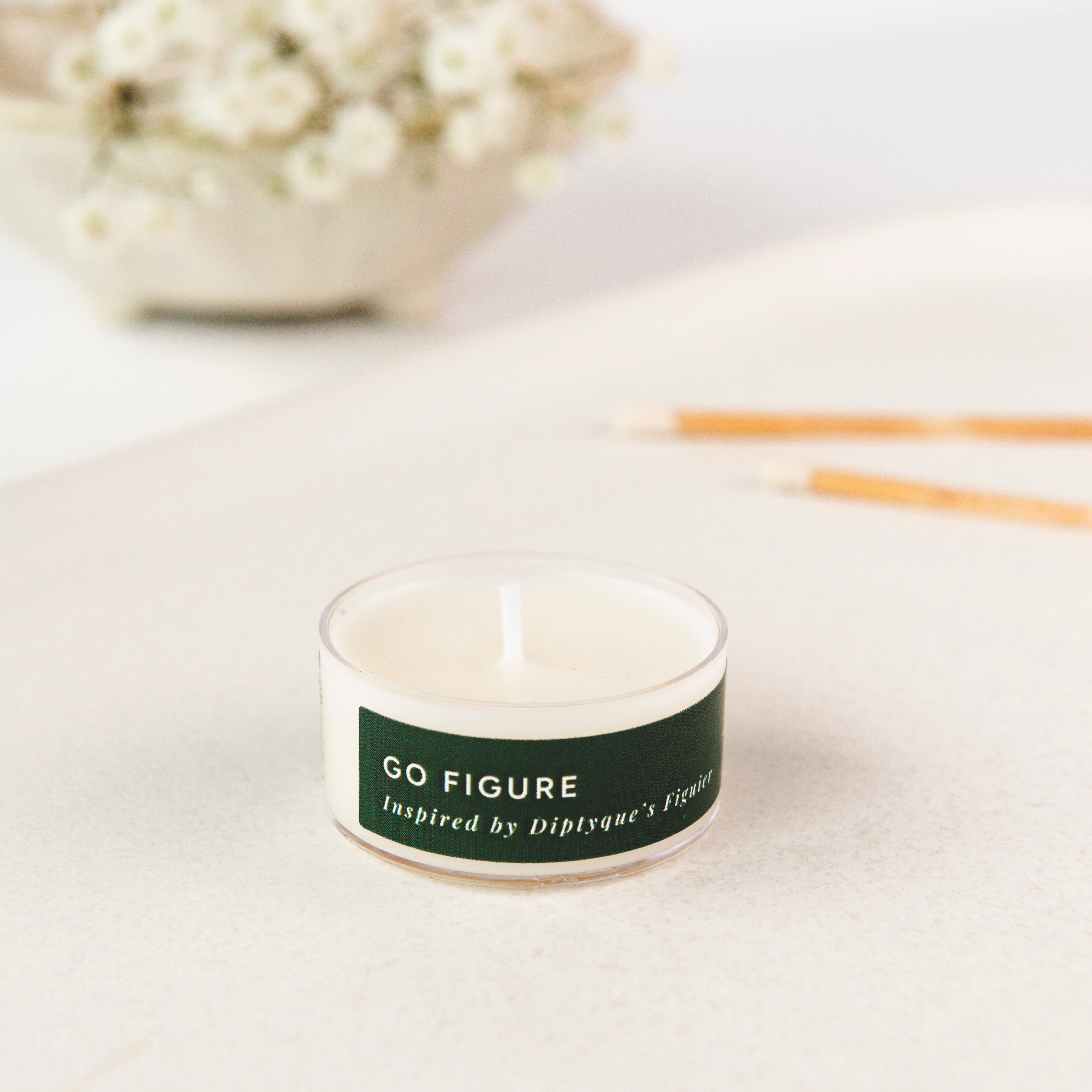Tea light candle with a label 'GO FIGURE' on a light surface with flowers in the background