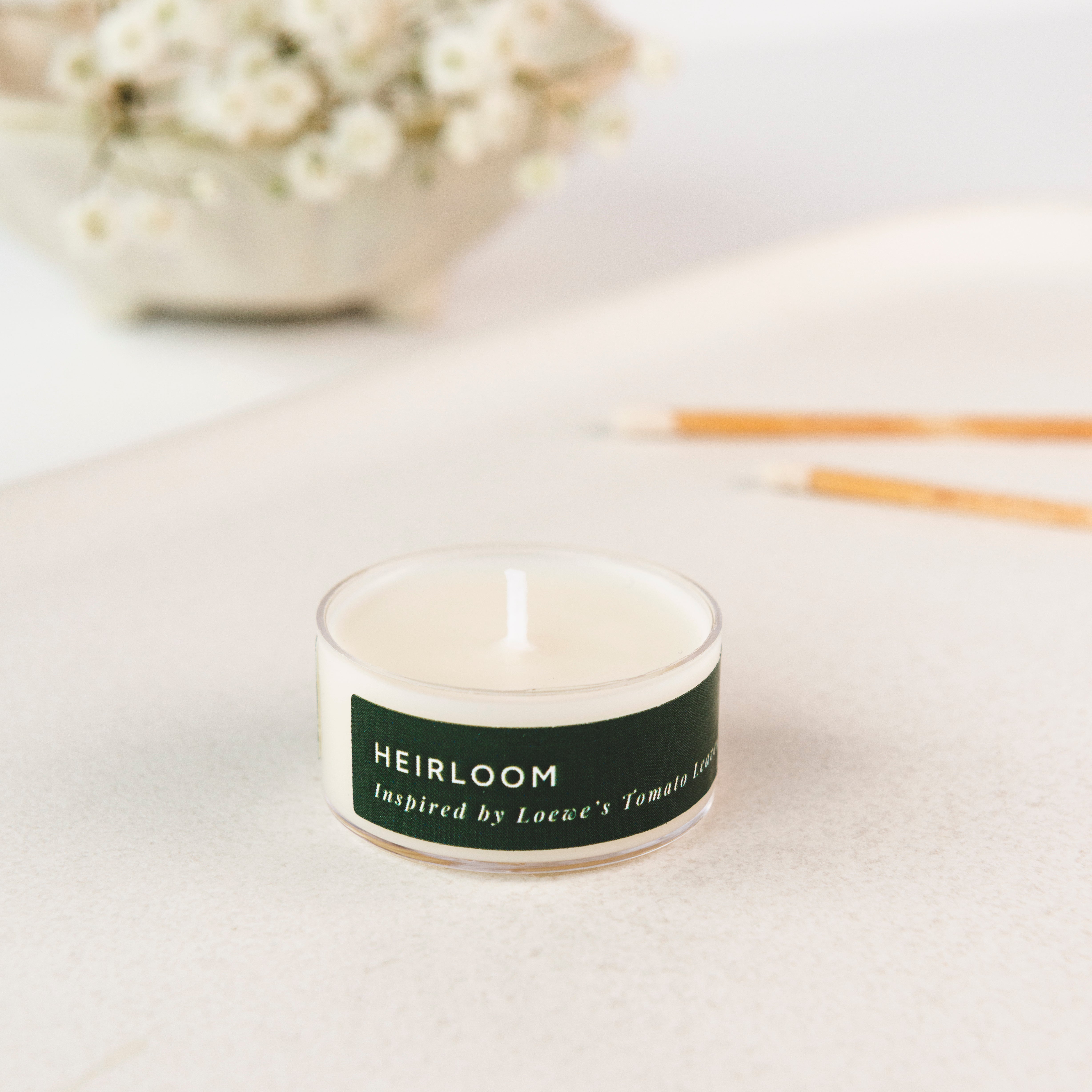 Tea light candle with 'Heirloom' label on a light surface with flowers in the background