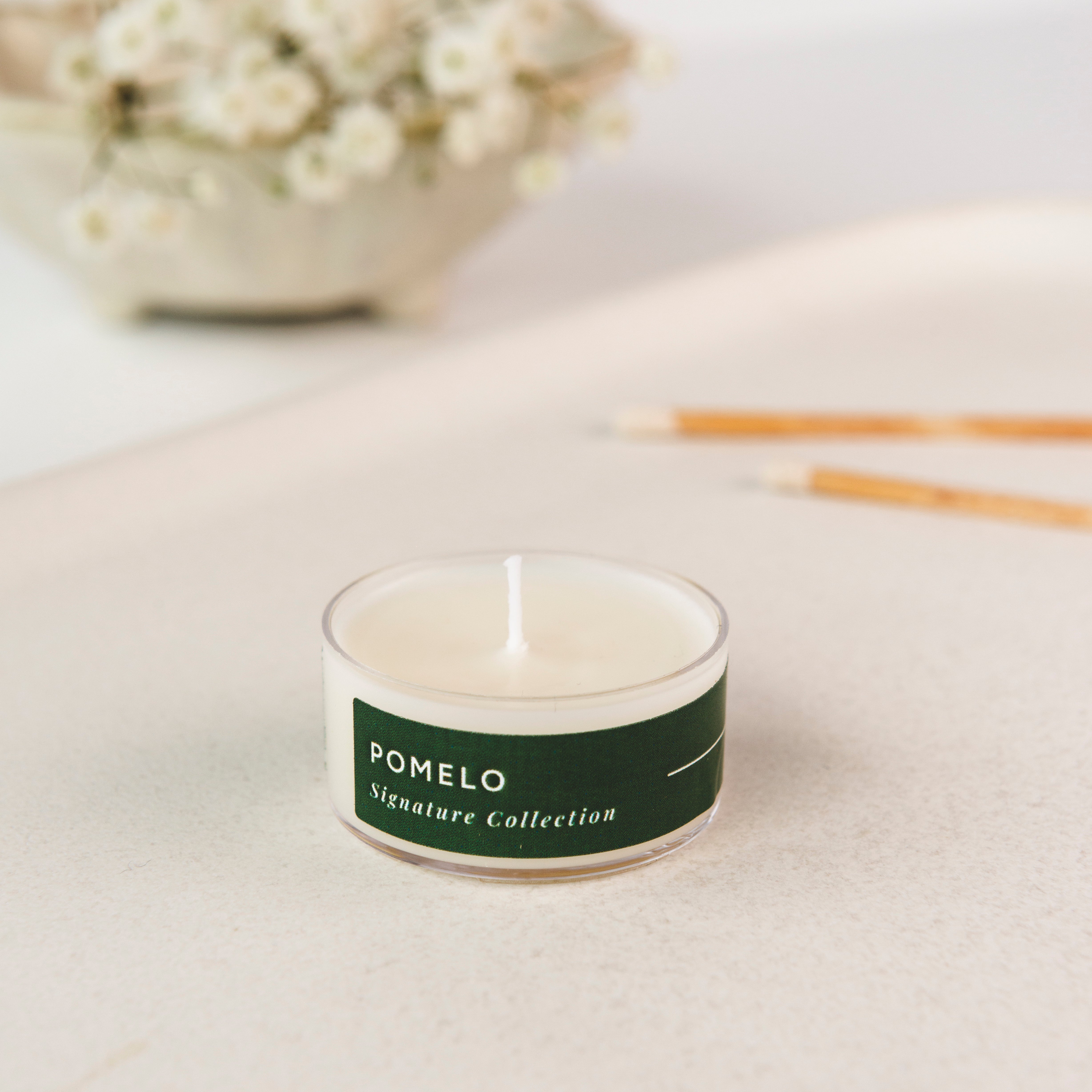 Tea light candle with 'Pomelo' label on a light surface with flowers and sticks in the background