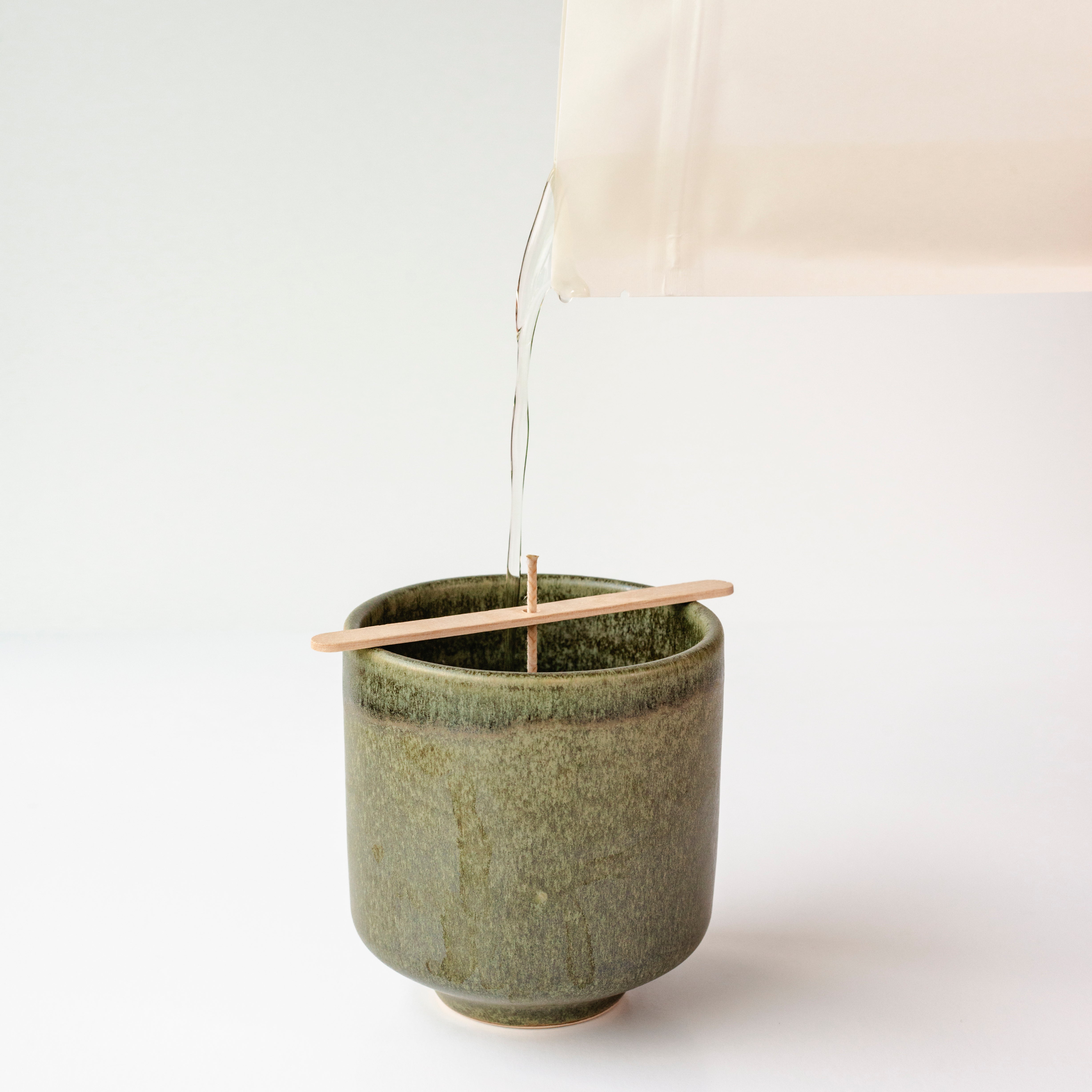 Candle wax being poured into a green ceramic vessel on a white background.