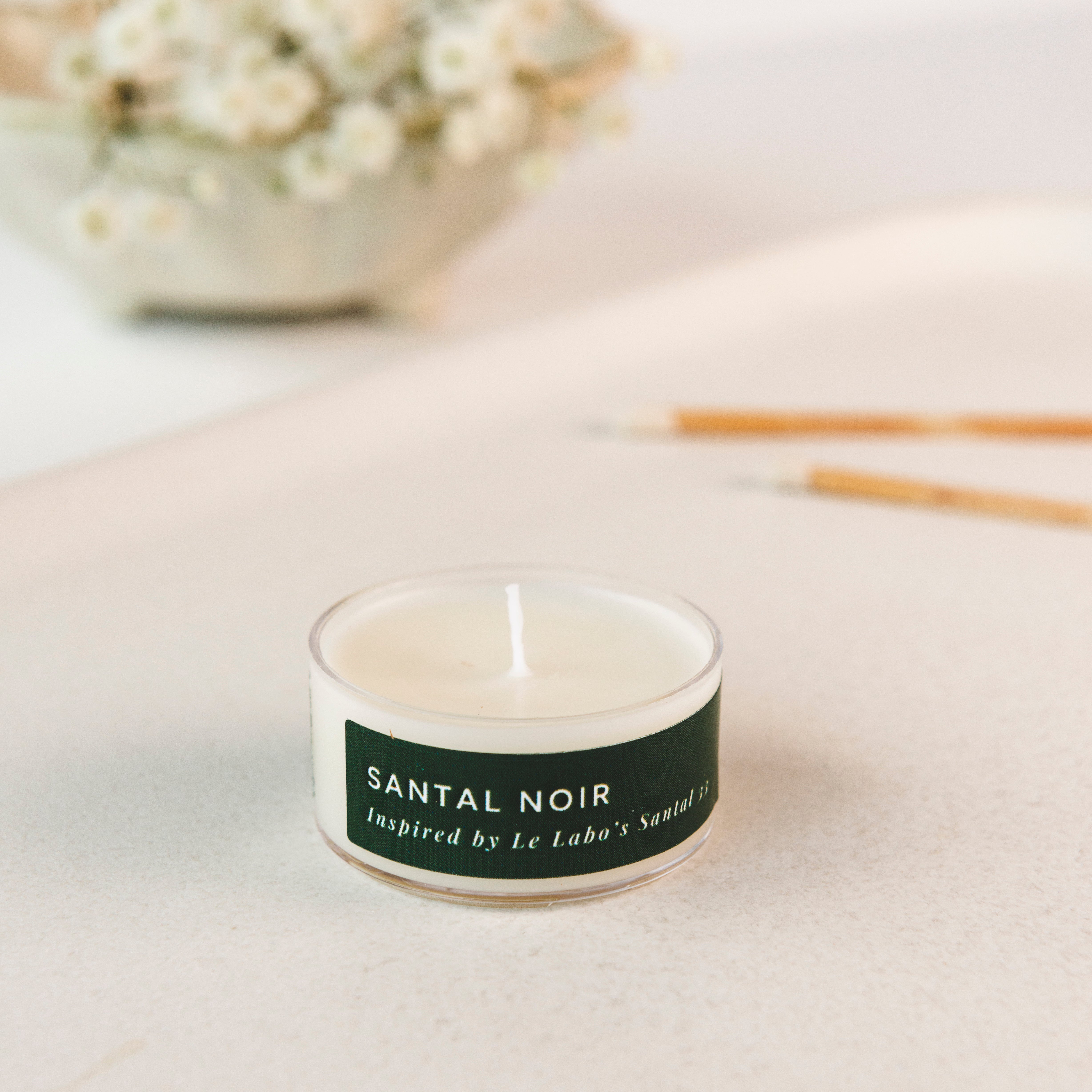 Tea light candle labeled 'Santal Noir' on a light surface with decorative elements in the background