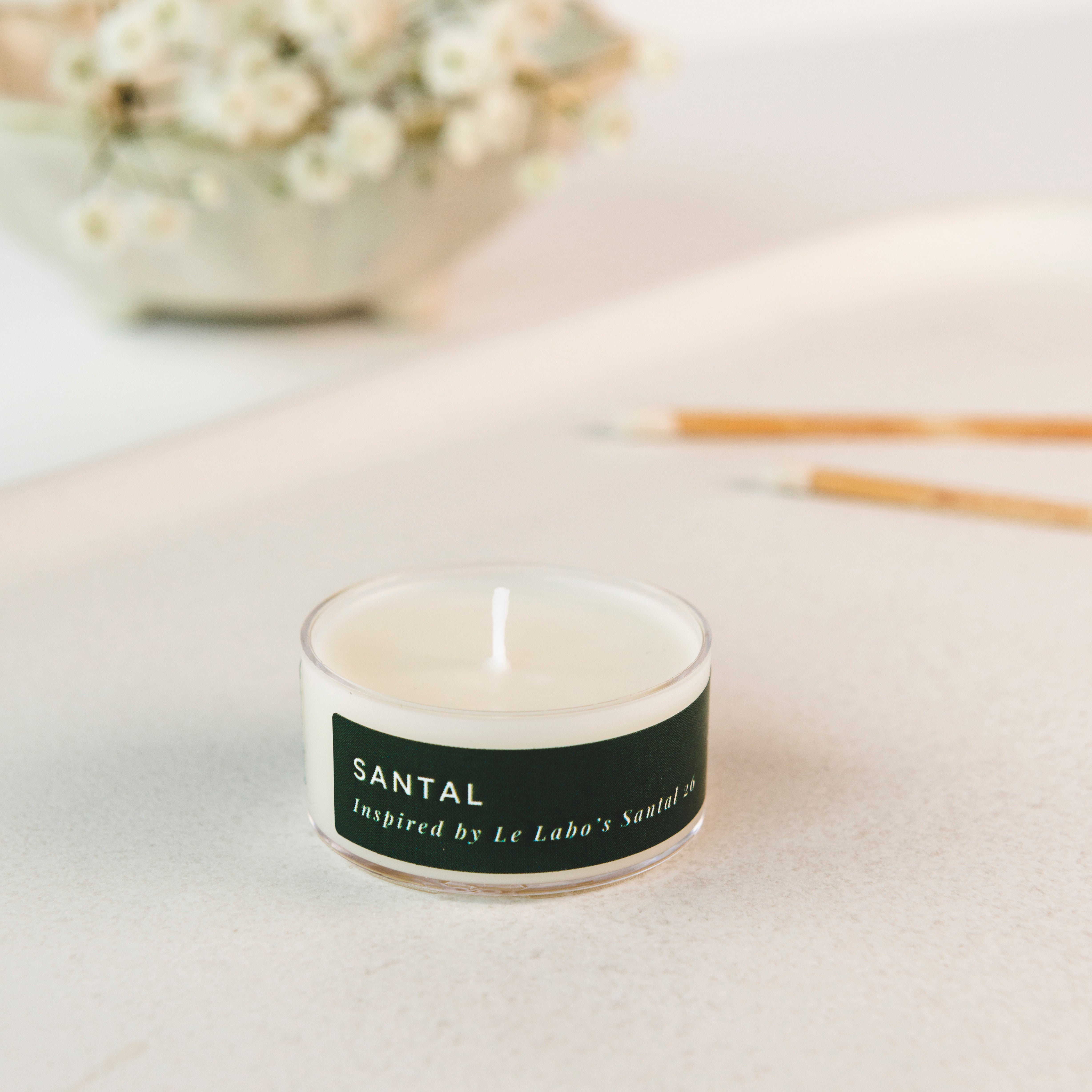 Tea light candle with a black label on a light surface, with flowers and sticks in the background