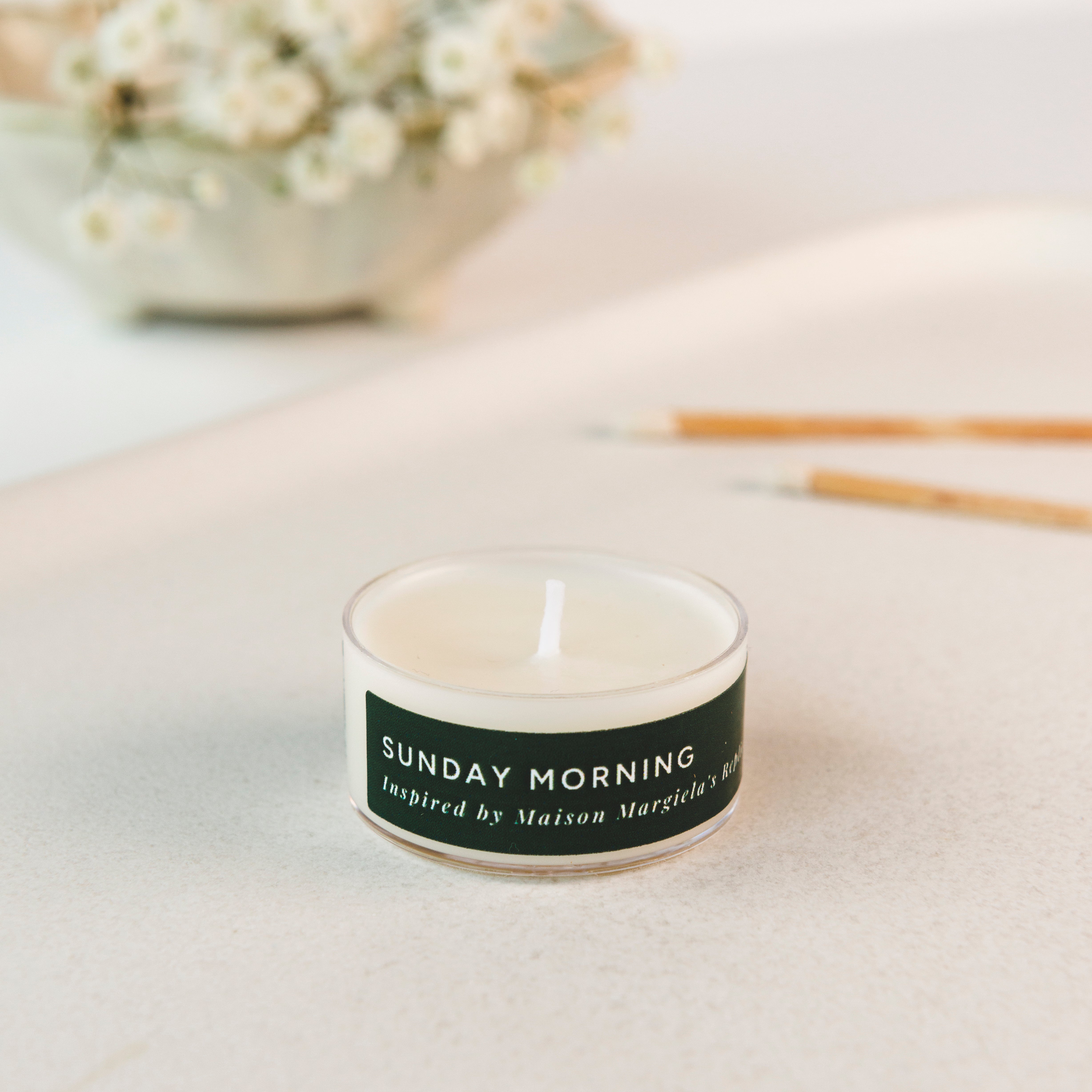 Tea light candle with a label reading 'Sunday Morning' on a light surface with flowers in the background