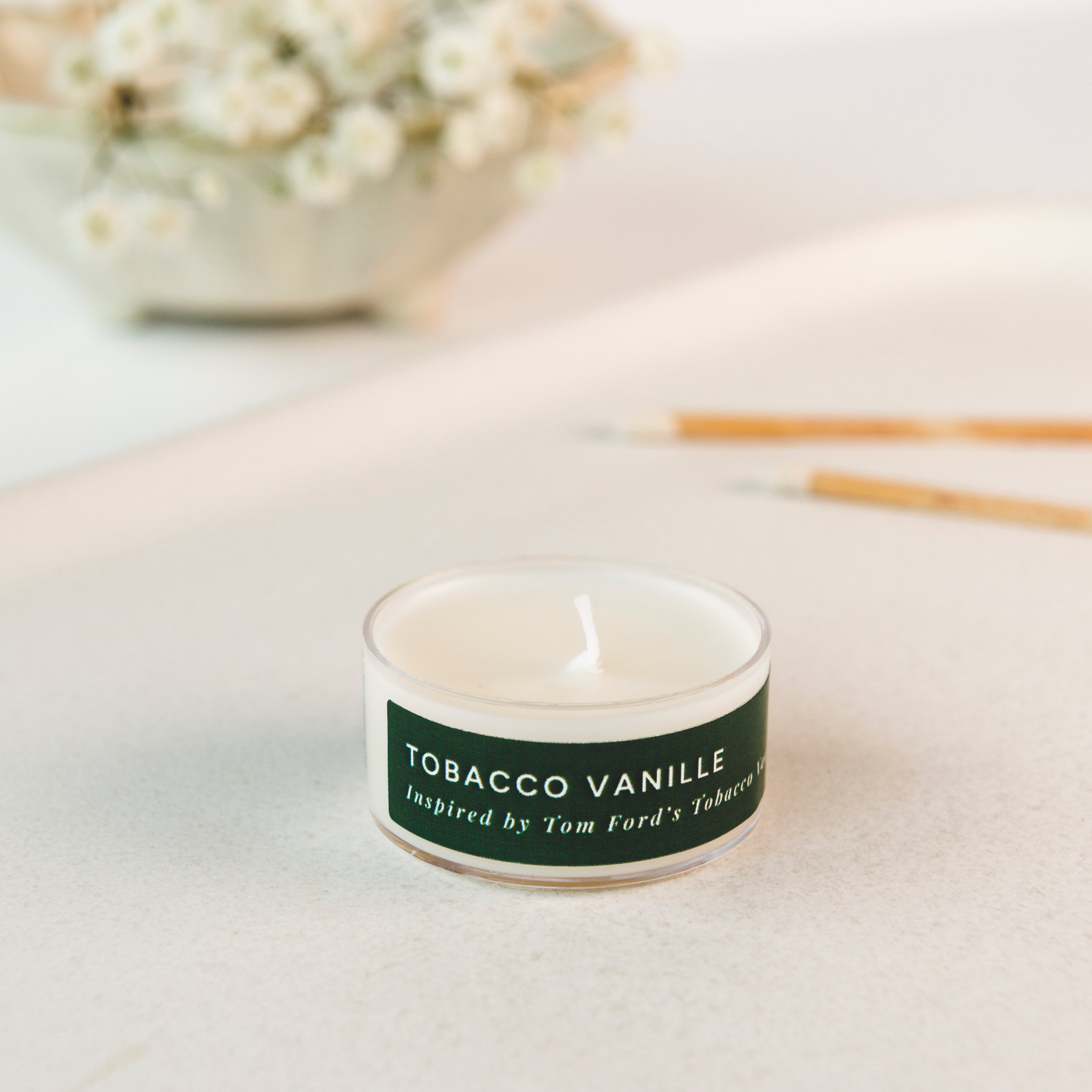 Tea light candle labeled 'Tobacco Vanille' on a light surface with decorative elements in the background