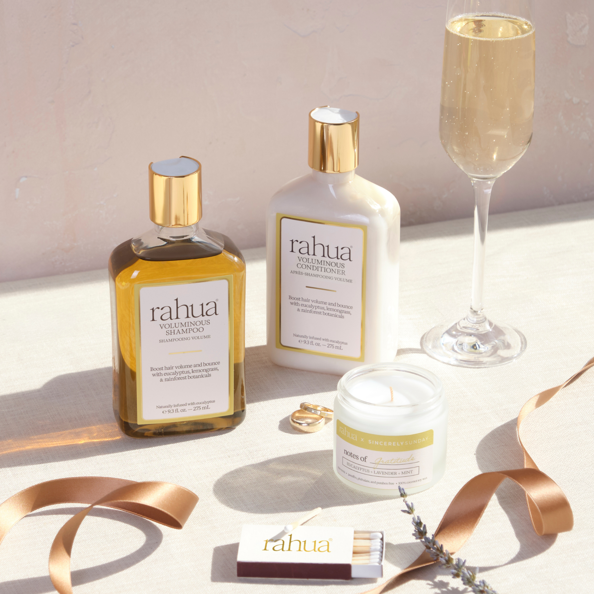 Rahua hair care products including shampoo, conditioner, and candle on a light surface with a glass of champagne.