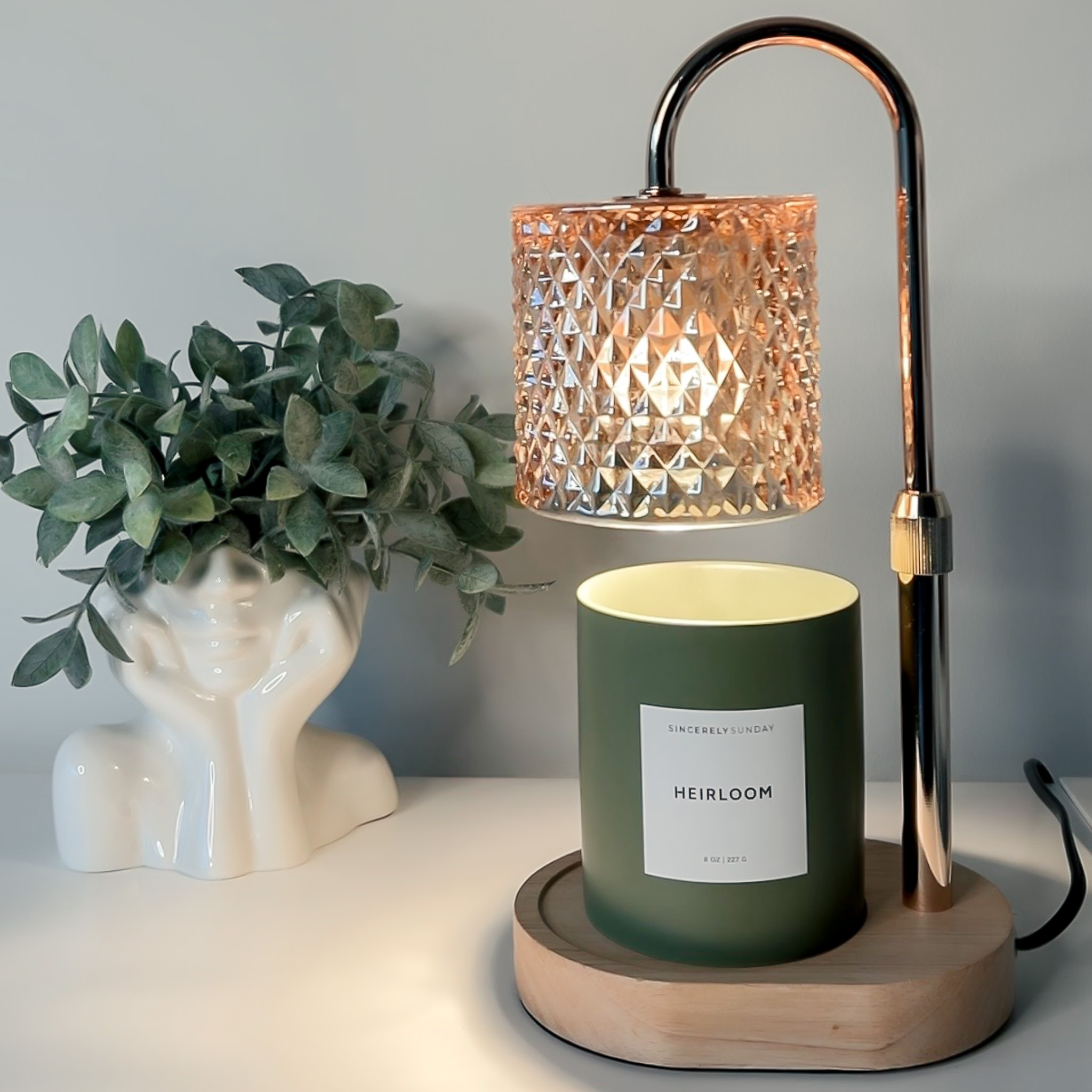 Candle labeled “Heirloom” under a heat lamp beside a plant on a white surface.