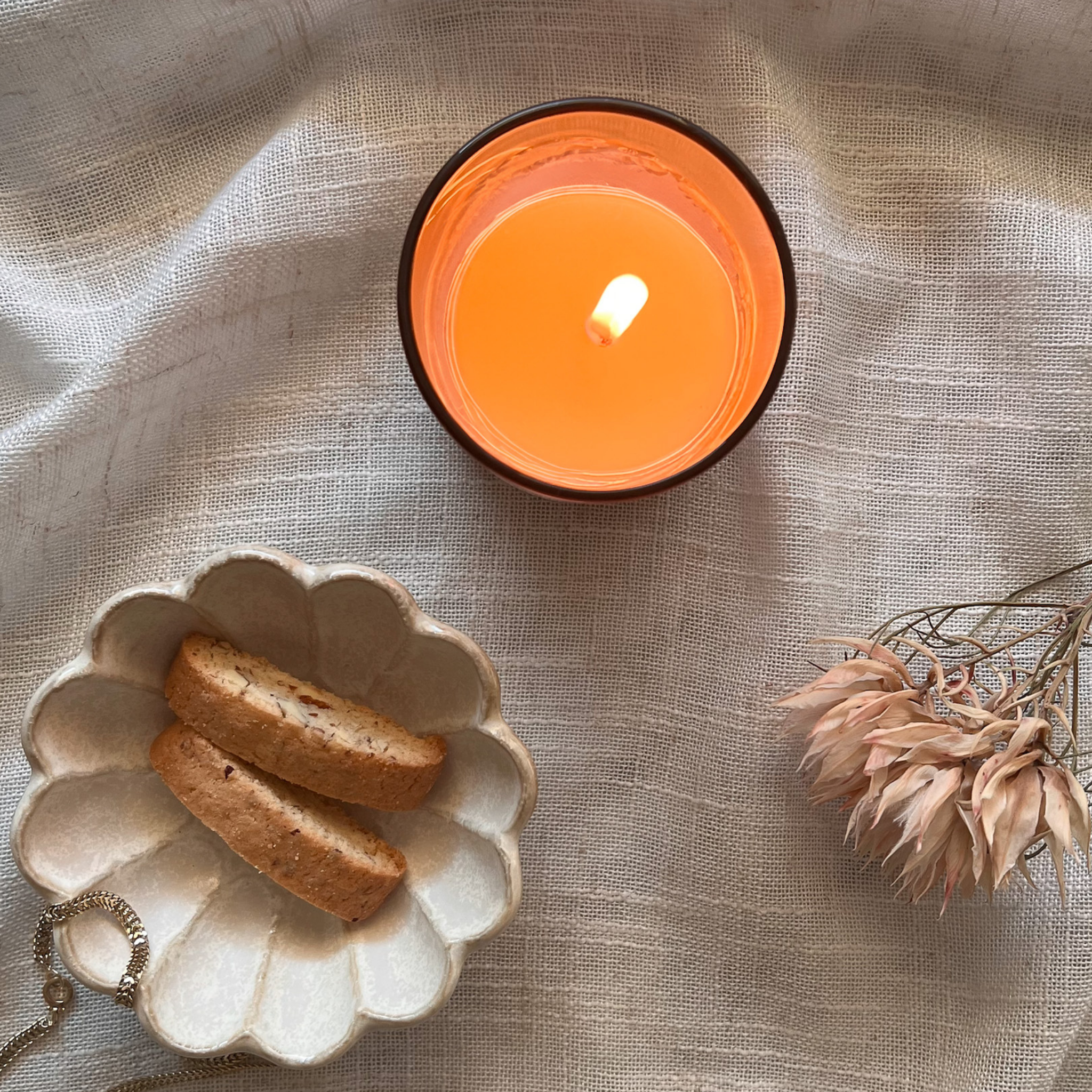 Candle with a small dish of cookies and a dried flower on a textured fabric background