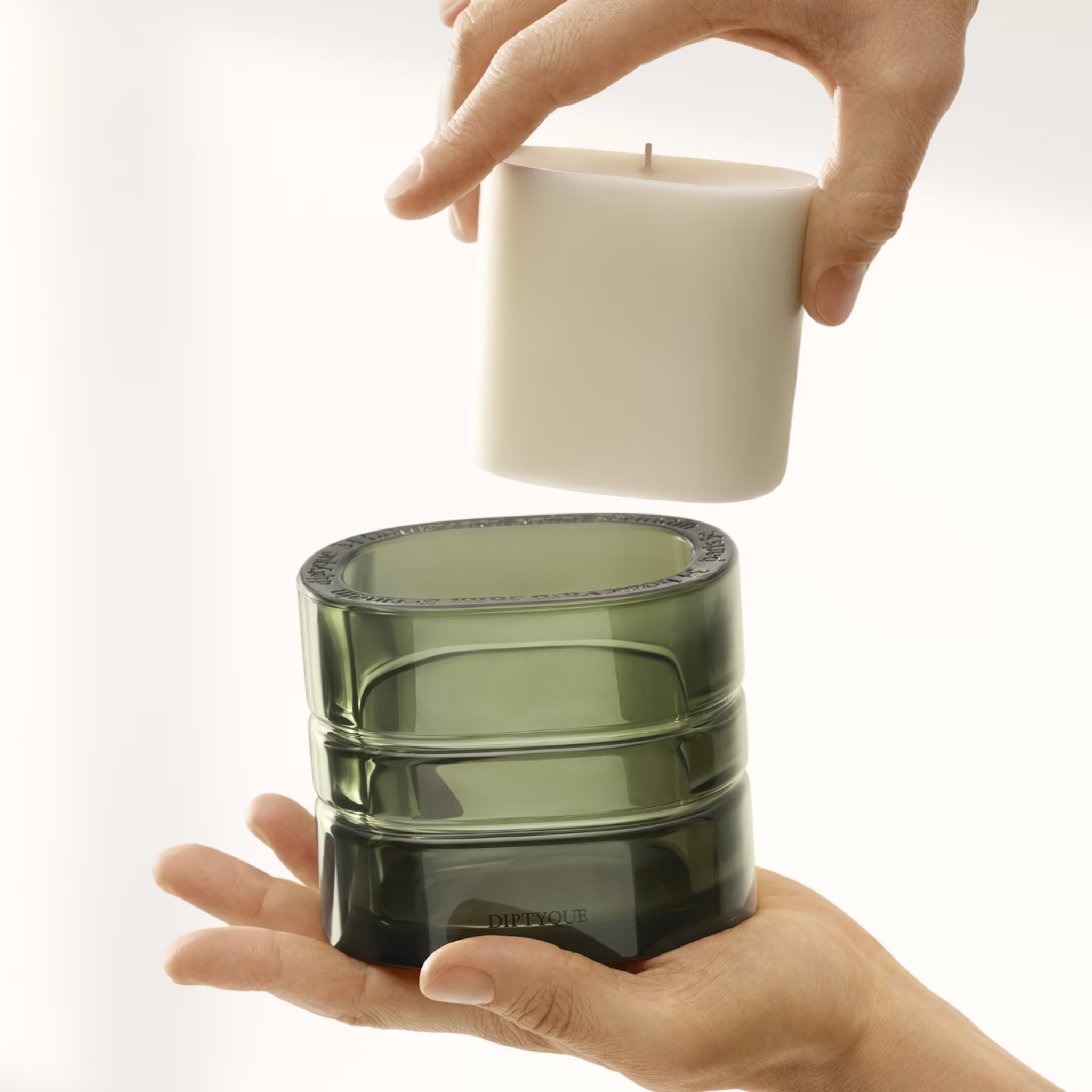 Hand holding a white candle refill insert above a green glass container on a white background.