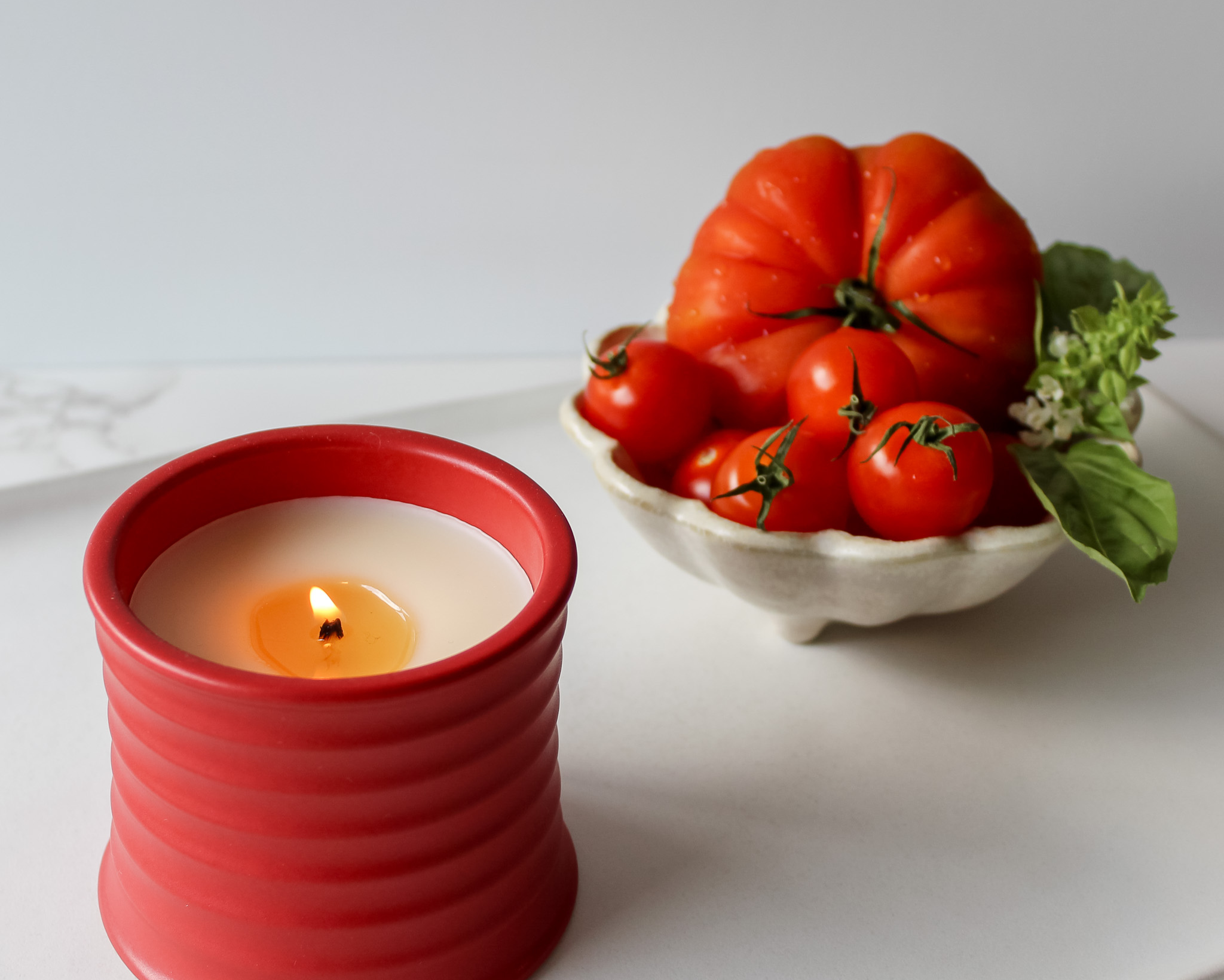 Red candle beside fresh tomatoes and basil in a scalloped dish on a white surface.