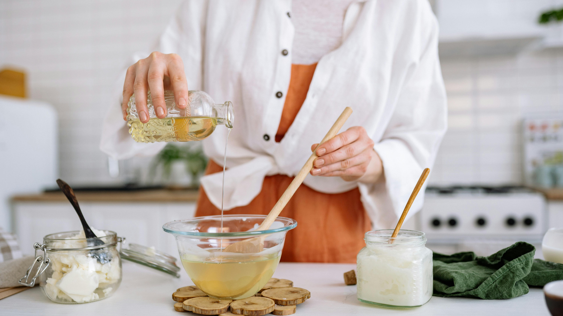 Person pouring fragrance oil into a glass bowl of melted wax on a kitchen counter.