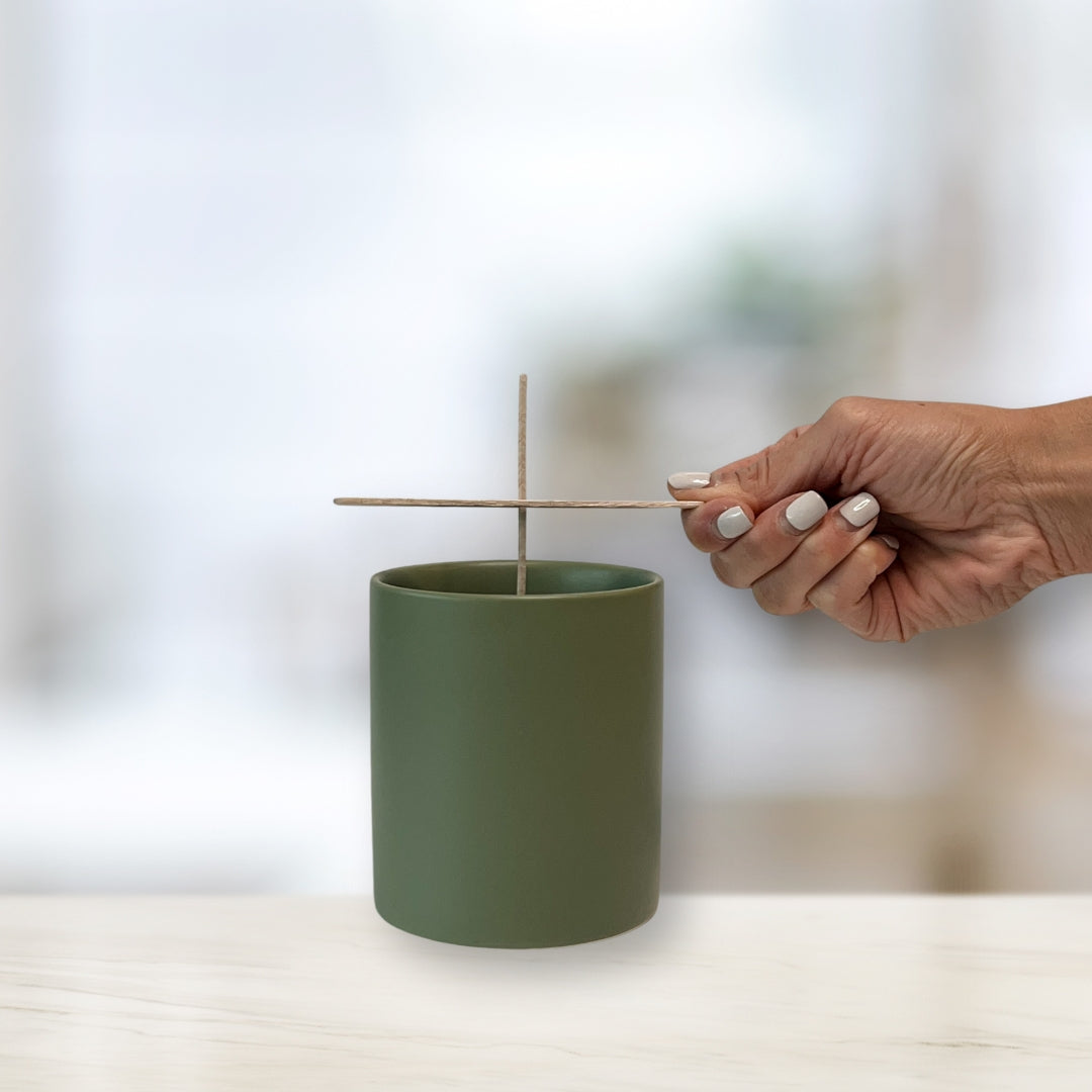Hand threading a cotton wick into a sage green candle vessel using a wick holder, preparing the vessel for candle making.