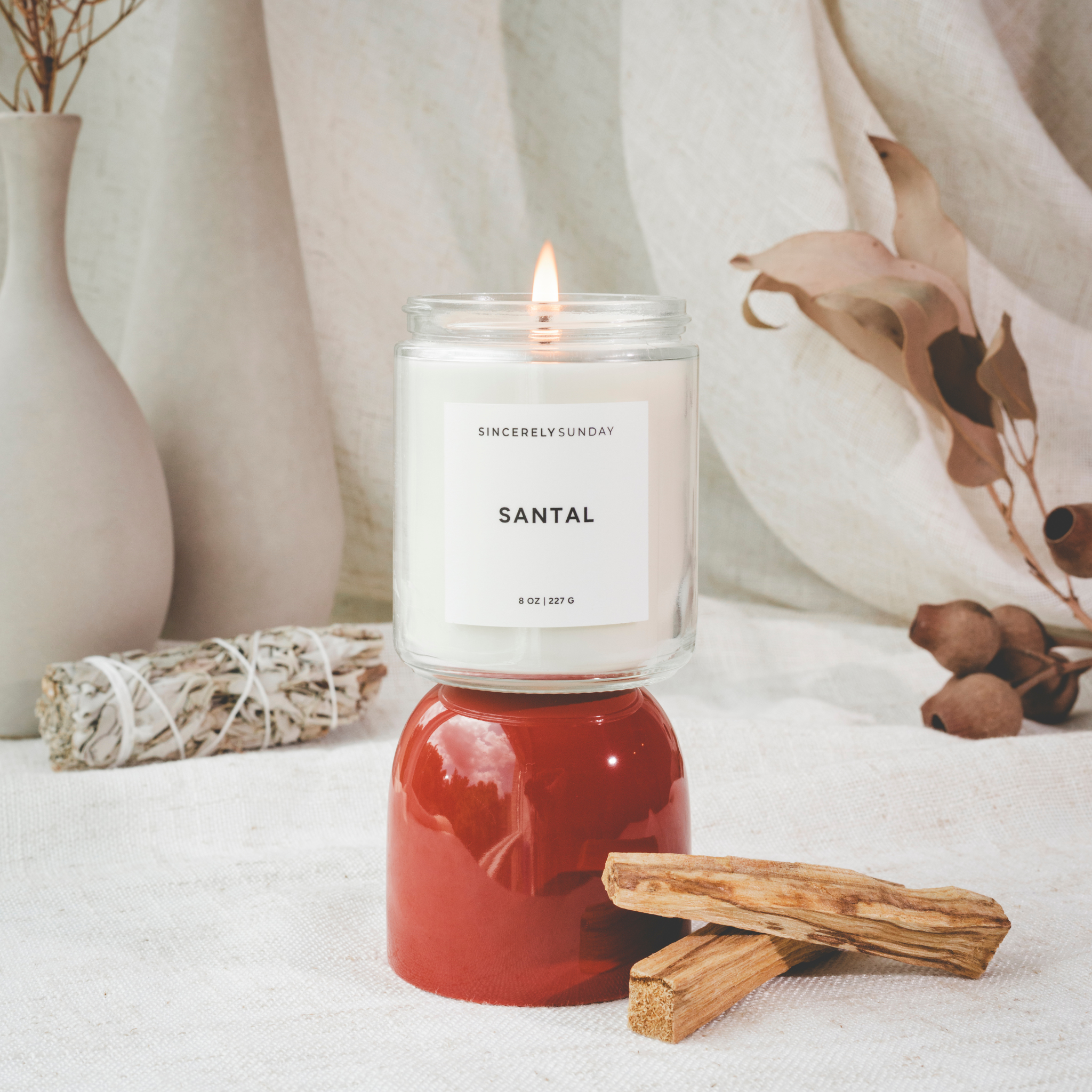 Candle with a red base labeled “Santal” on a white surface, styled with palo santo sticks, sage, and a vase.