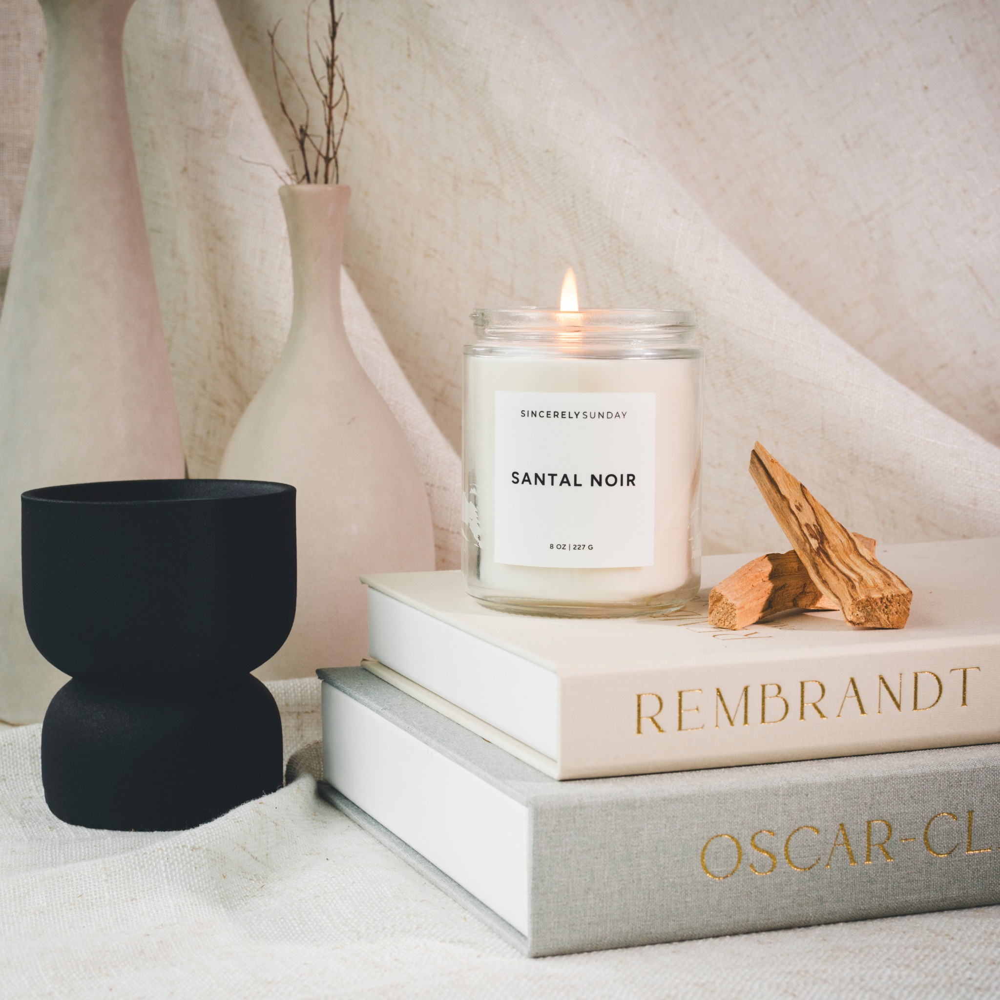 Candle labeled “Santal Noir” on books beside a black vessel and palo santo in a decorative setup.