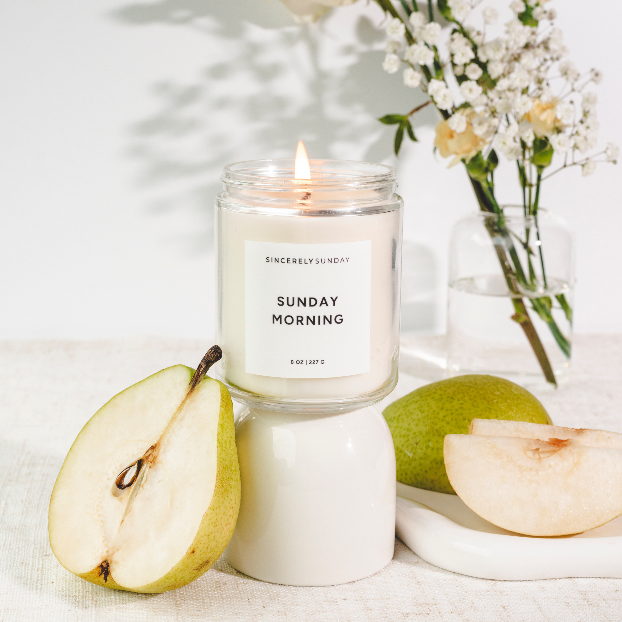 Candle labeled “Sunday Morning” on a white base with fresh pears and white flowers on a light surface.