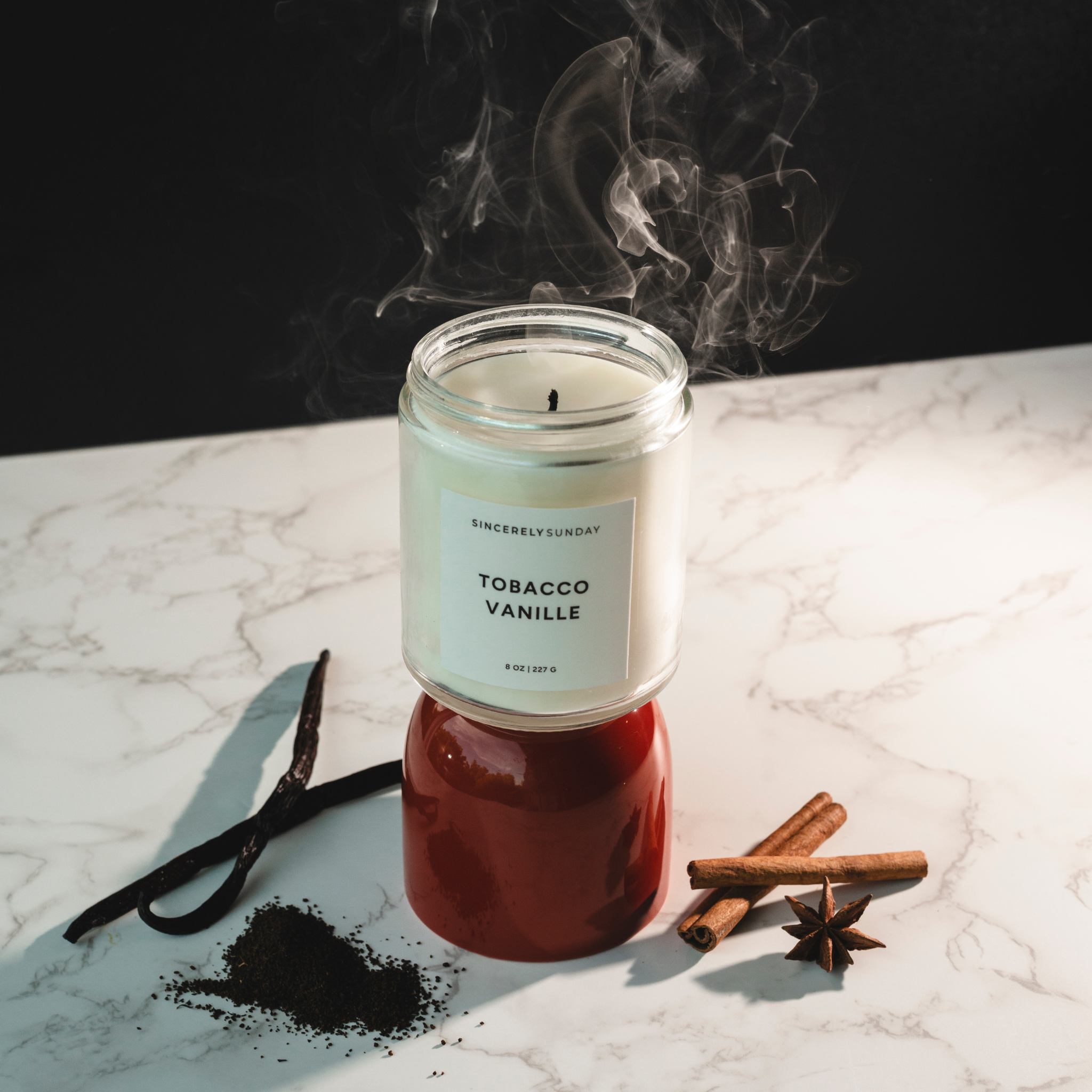 Candle labeled “Tobacco Vanille” with vanilla beans, cinnamon sticks, and assorted spices on a marble surface against a dark background.