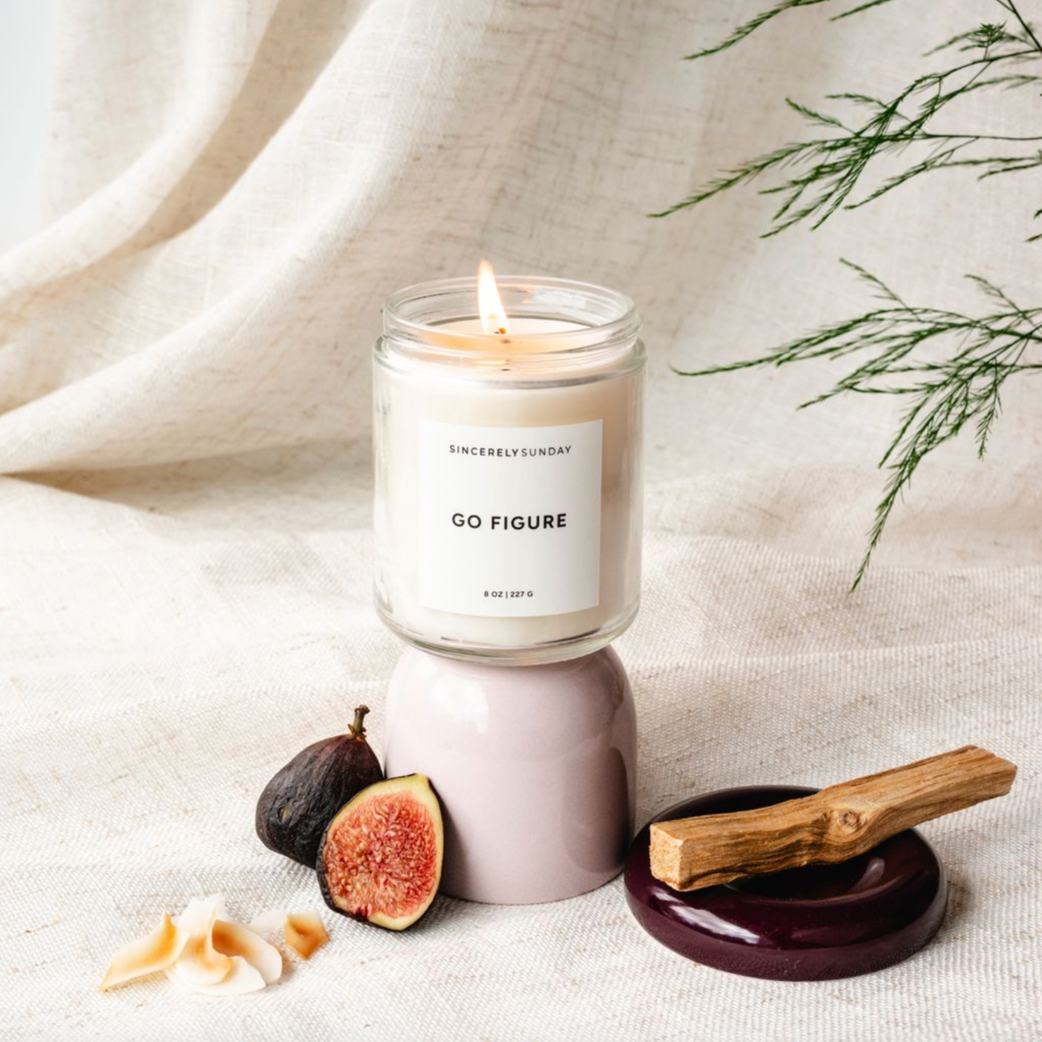 Candle in a glass jar labeled “Go Figure” with palo santo, fresh figs, and coconut flakes on a neutral background.