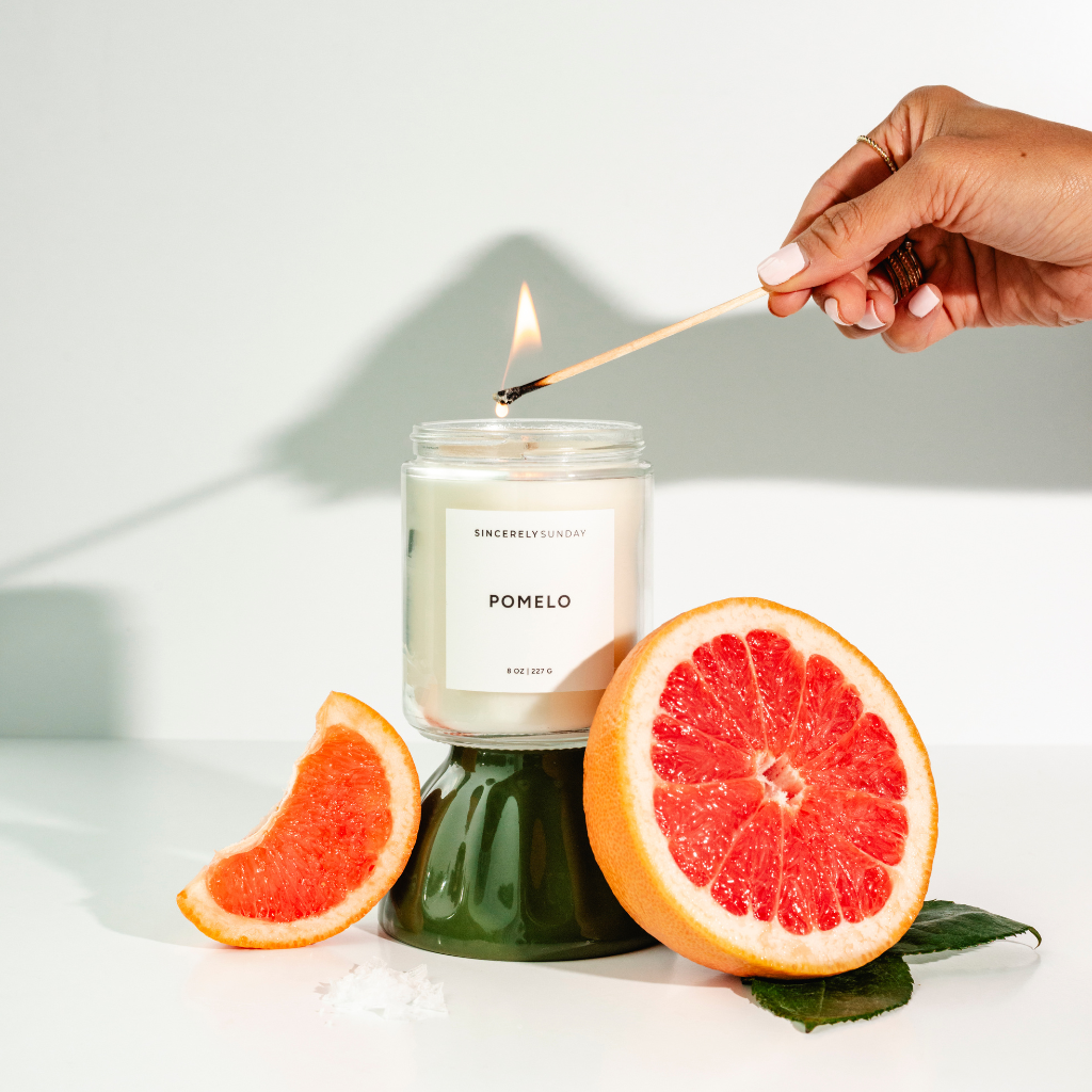 Candle labeled 'Pomelo' being lit with a match, surrounded by grapefruit slices on a white background.