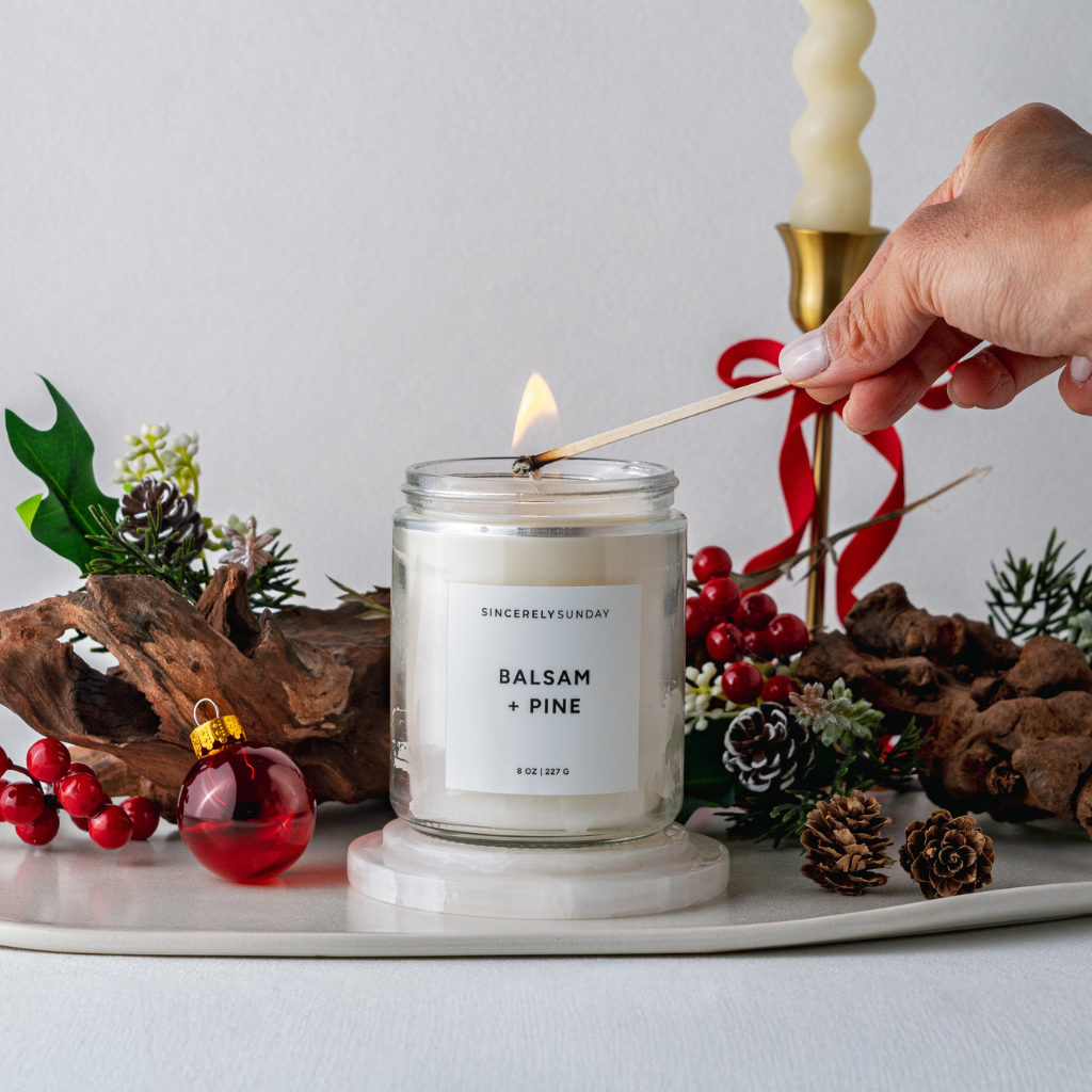 Candle labeled 'Balsam + Pine' being lit with a match, surrounded by festive decorations.