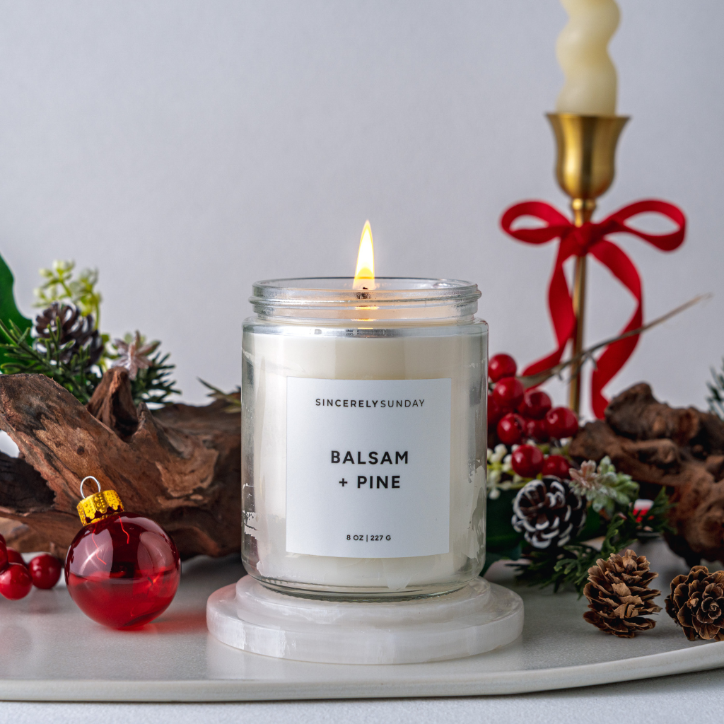Candle labeled 'Balsam + Pine' with Christmas decorations on a white surface