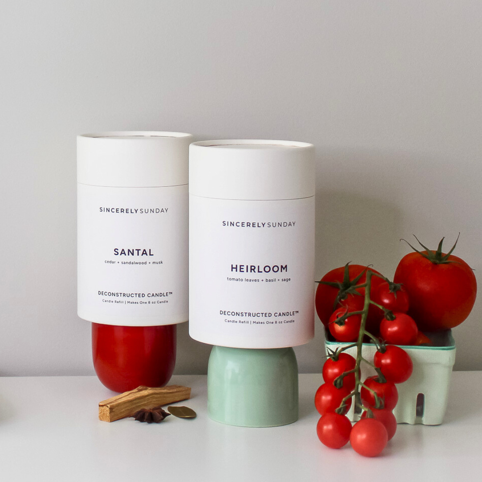 Two candles labeled 'Santal' and 'Heirloom' with tomatoes and palo santo sticks on a white surface.