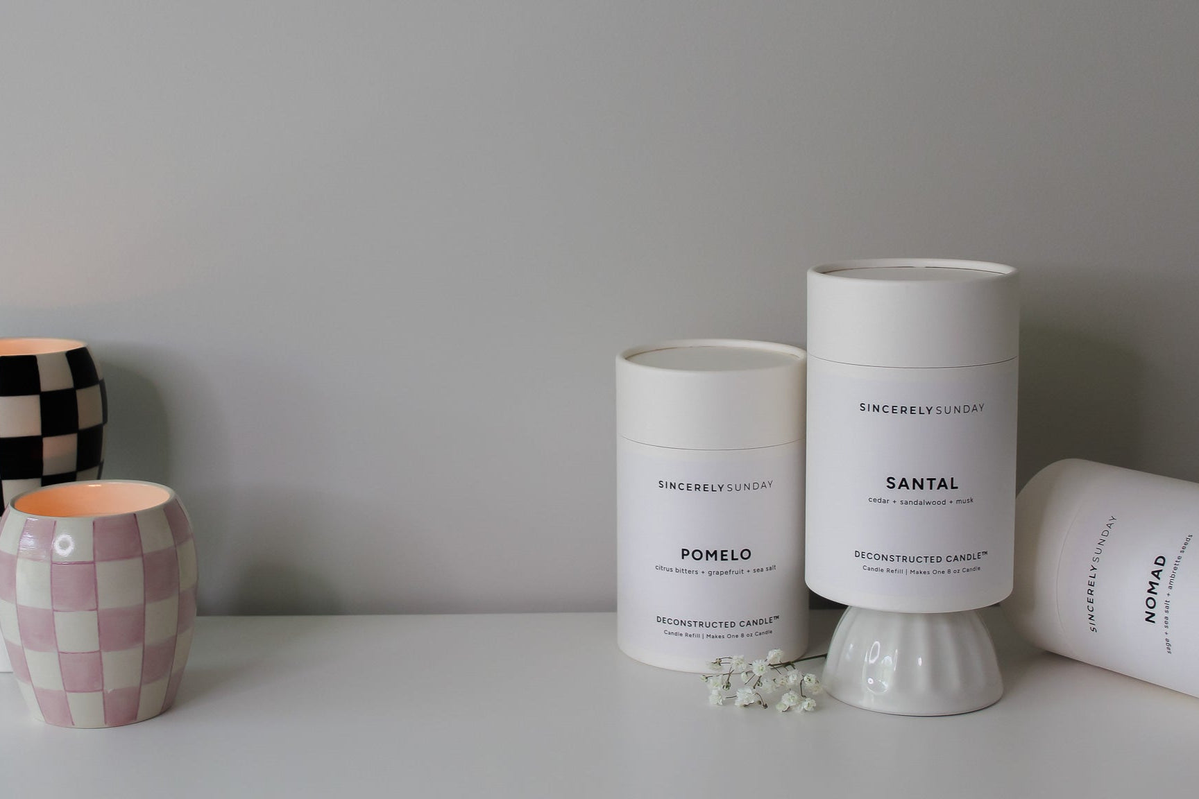 Three cylindrical candle refill kits next to two candles on a surface against a gray wall