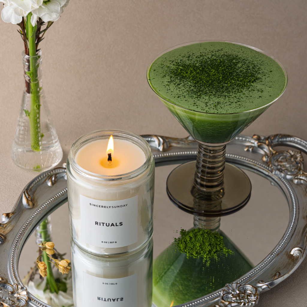 Candle labeled 'Rituals' next to a green drink in a martini glass on a reflective surface.