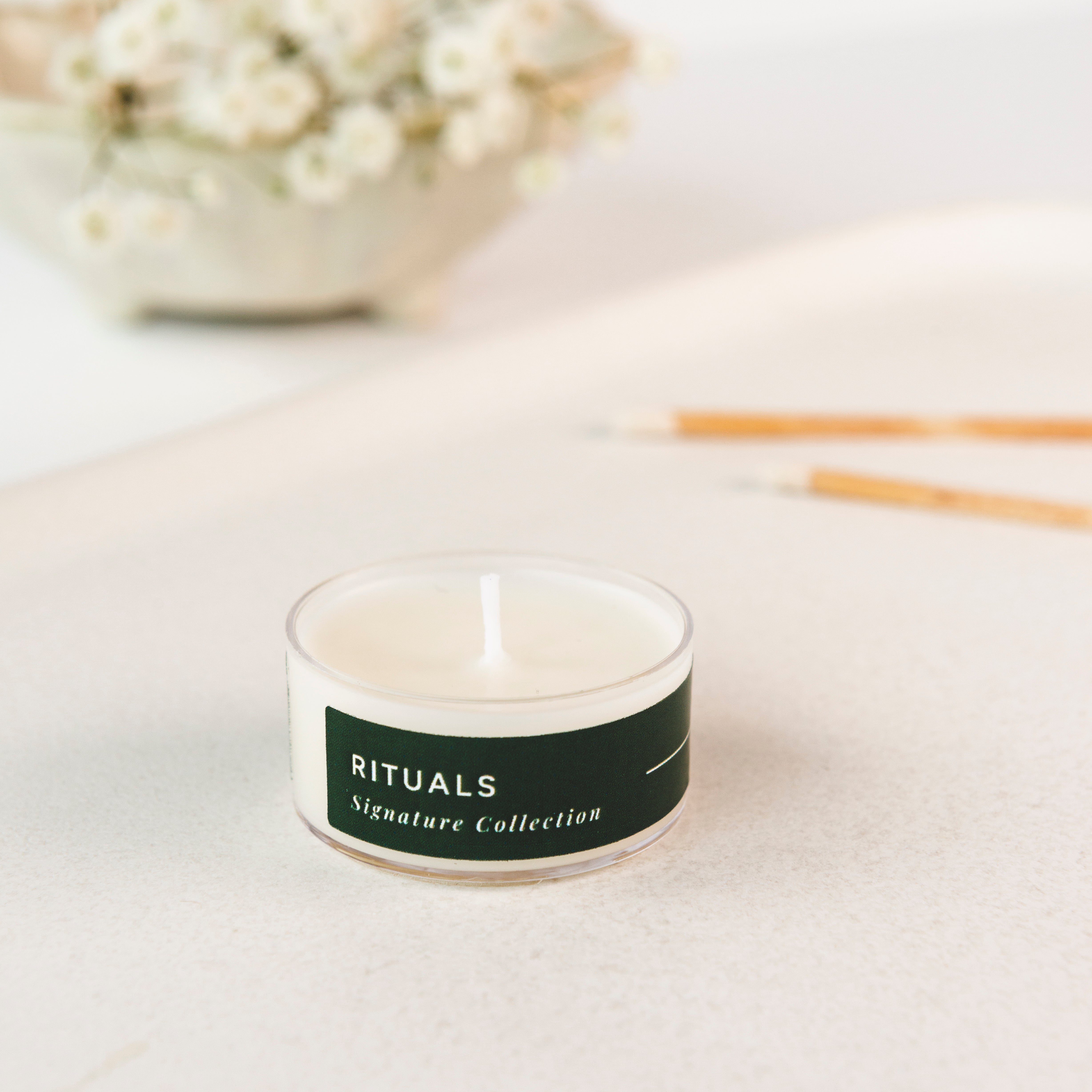 Tea light candle with 'Rituals' label on a light surface with flowers and sticks in the background