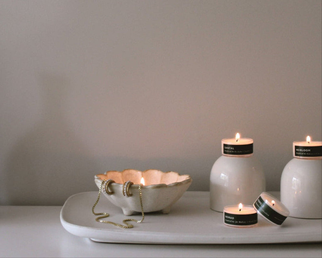 Four lit tea light candles on a tray beside a vase and scalloped ceramic dish with jewelry draped over it. 