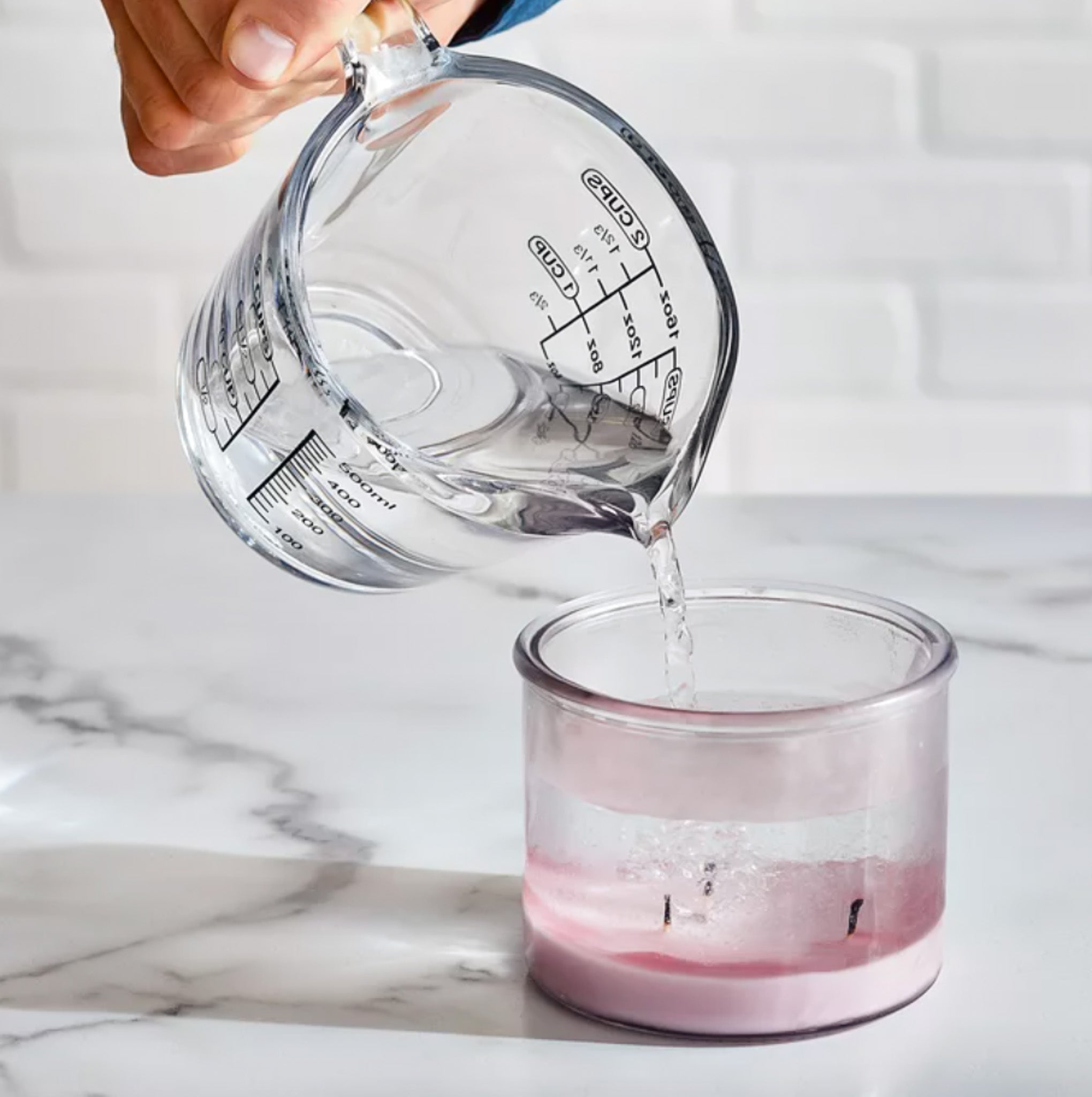 Measuring cup pouring hot water into a glass candle jar with leftover wax.