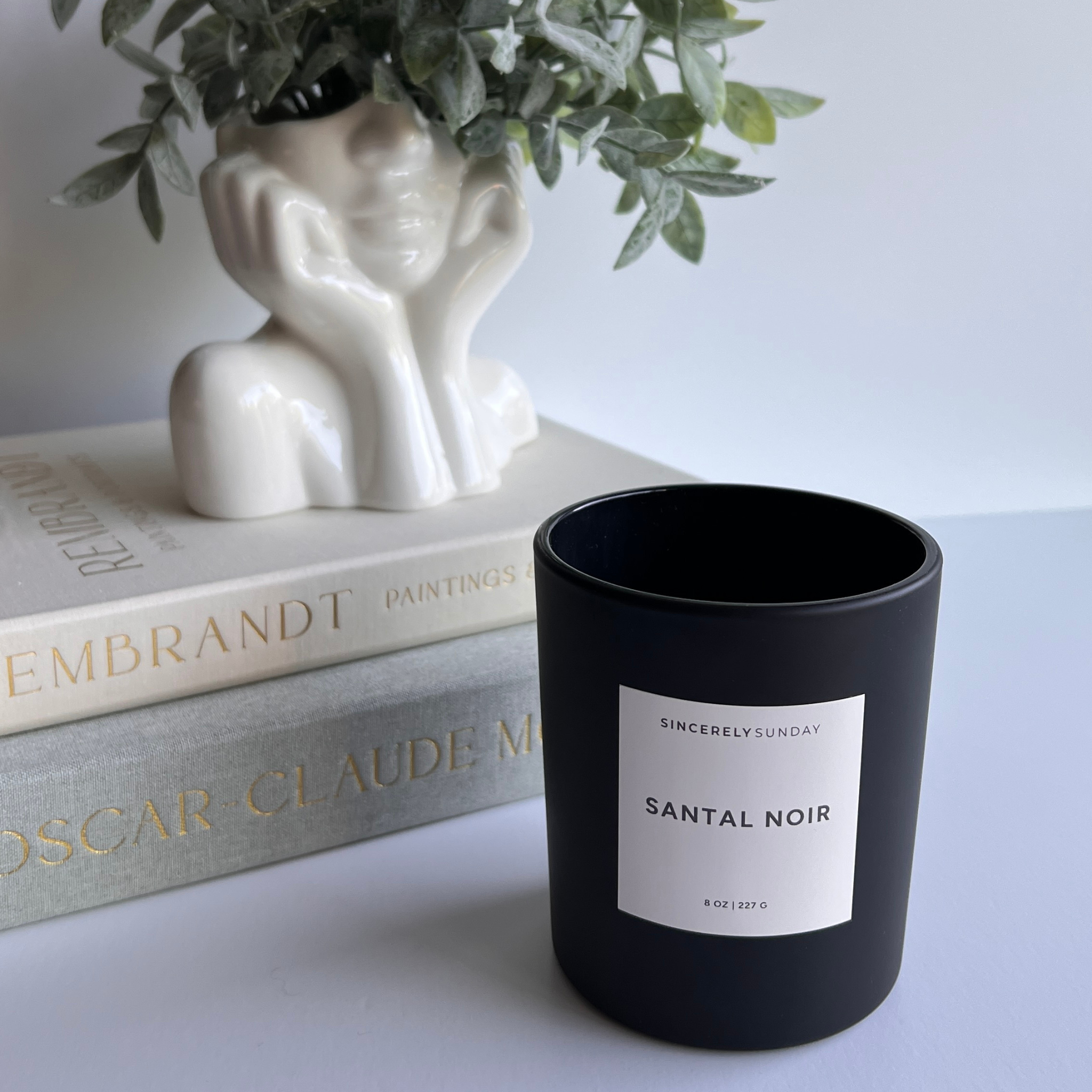 Black candle labeled 'Santal Noir' on a surface with books and a decorative vase in the background.