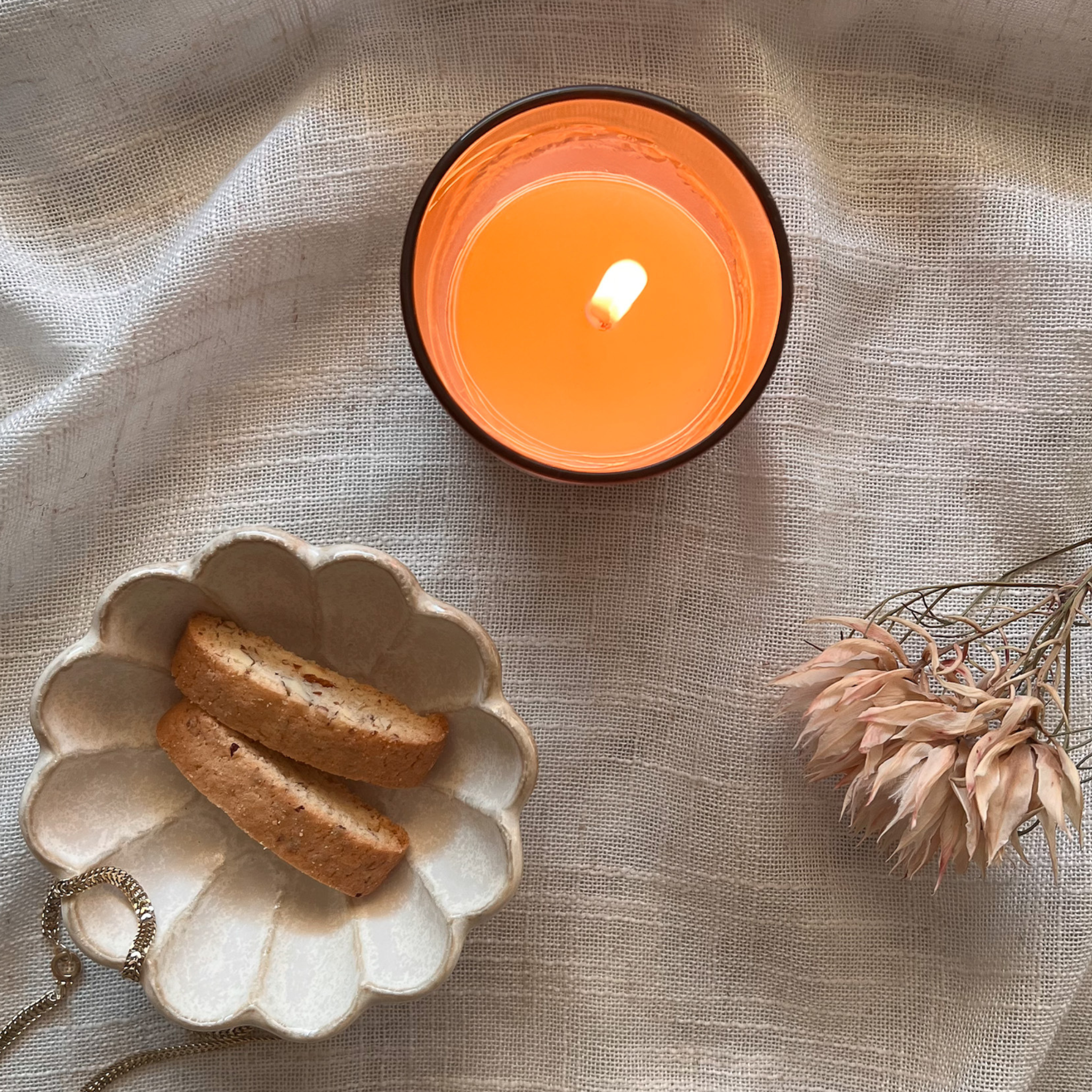 Candle burning beside a scalloped ceramic bowl of cookies and a dried flower on a textured surface.
