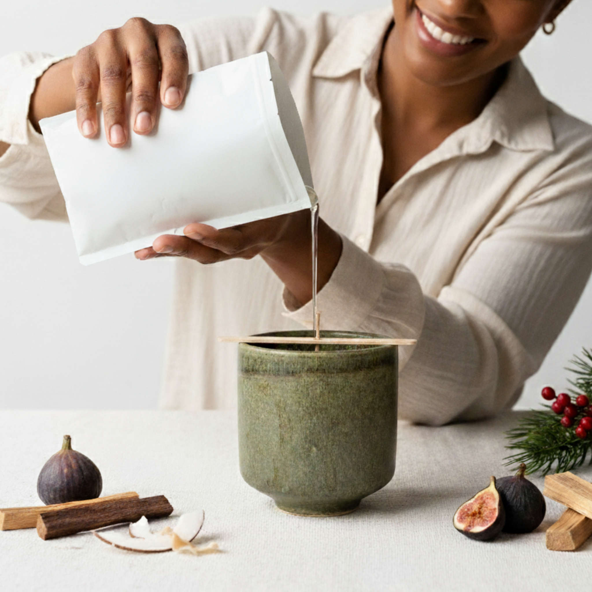 Person pouring liquid from a white pouch into a green ceramic vessel on a light surface with figs and palo santo