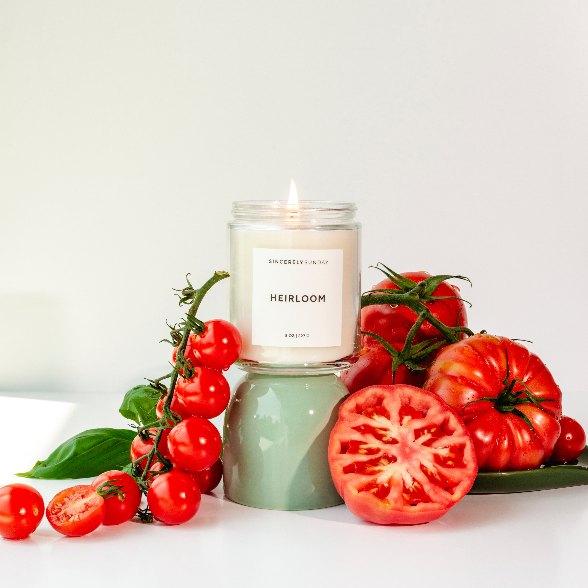 Candle labeled 'Heirloom' surrounded by fresh tomatoes on a white background