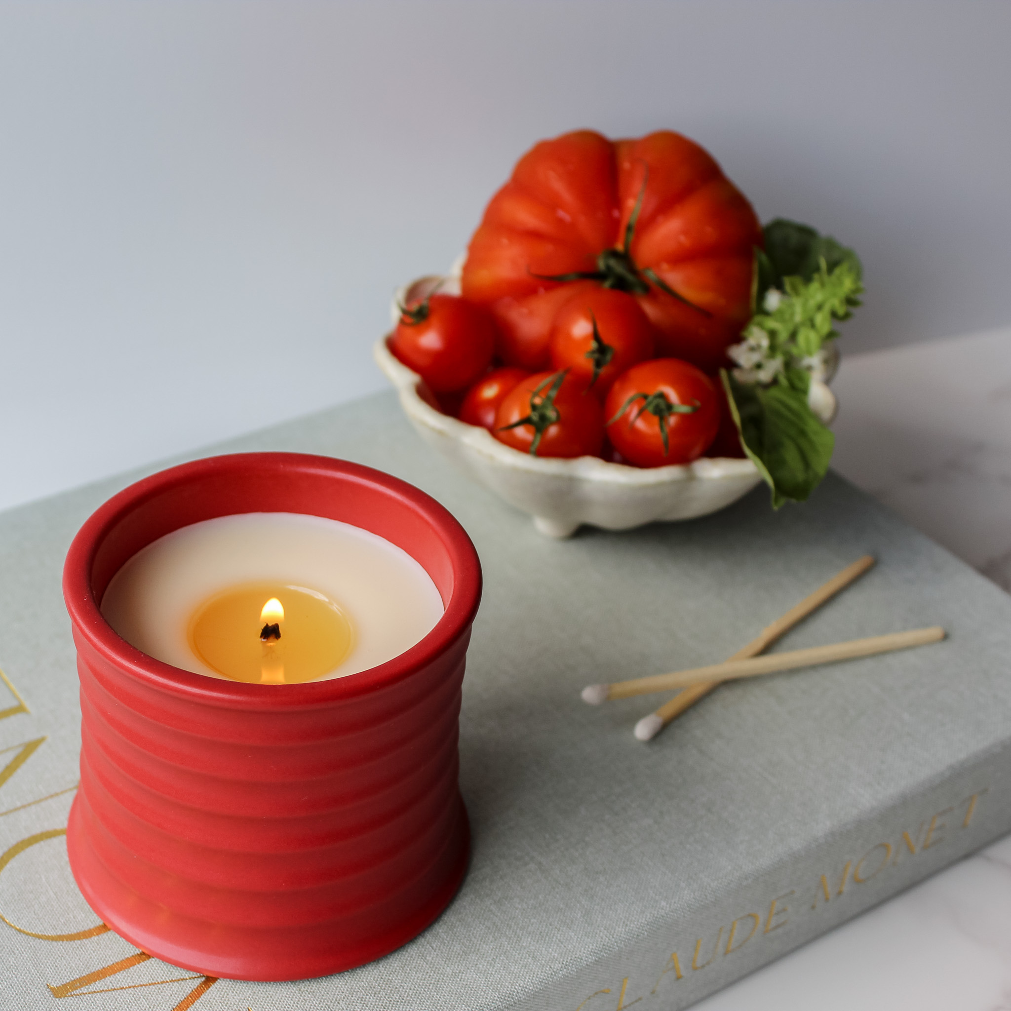 Red candle on a white tray next to a scalloped dish filled with fresh tomatoes and basil on a white background.