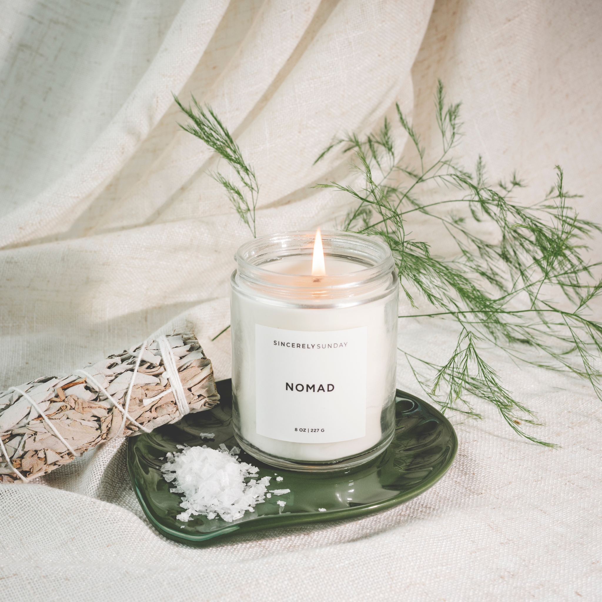 Candle labeled “Nomad” on a green plate with sage and sea salt, styled on a light background.