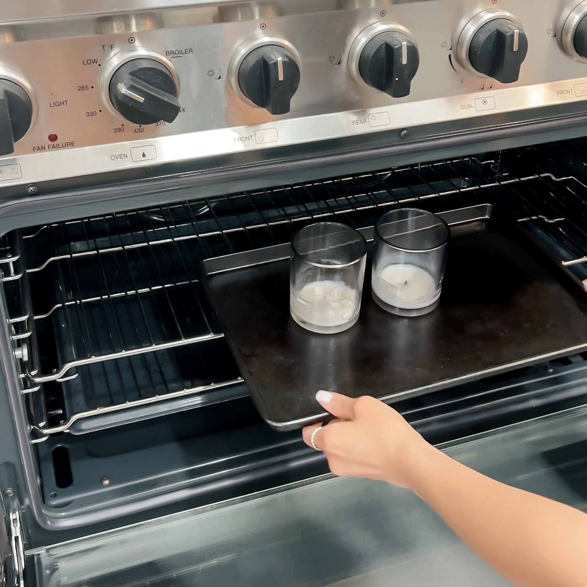 Person placing two glass candles with leftover wax on a baking sheet inside an oven.