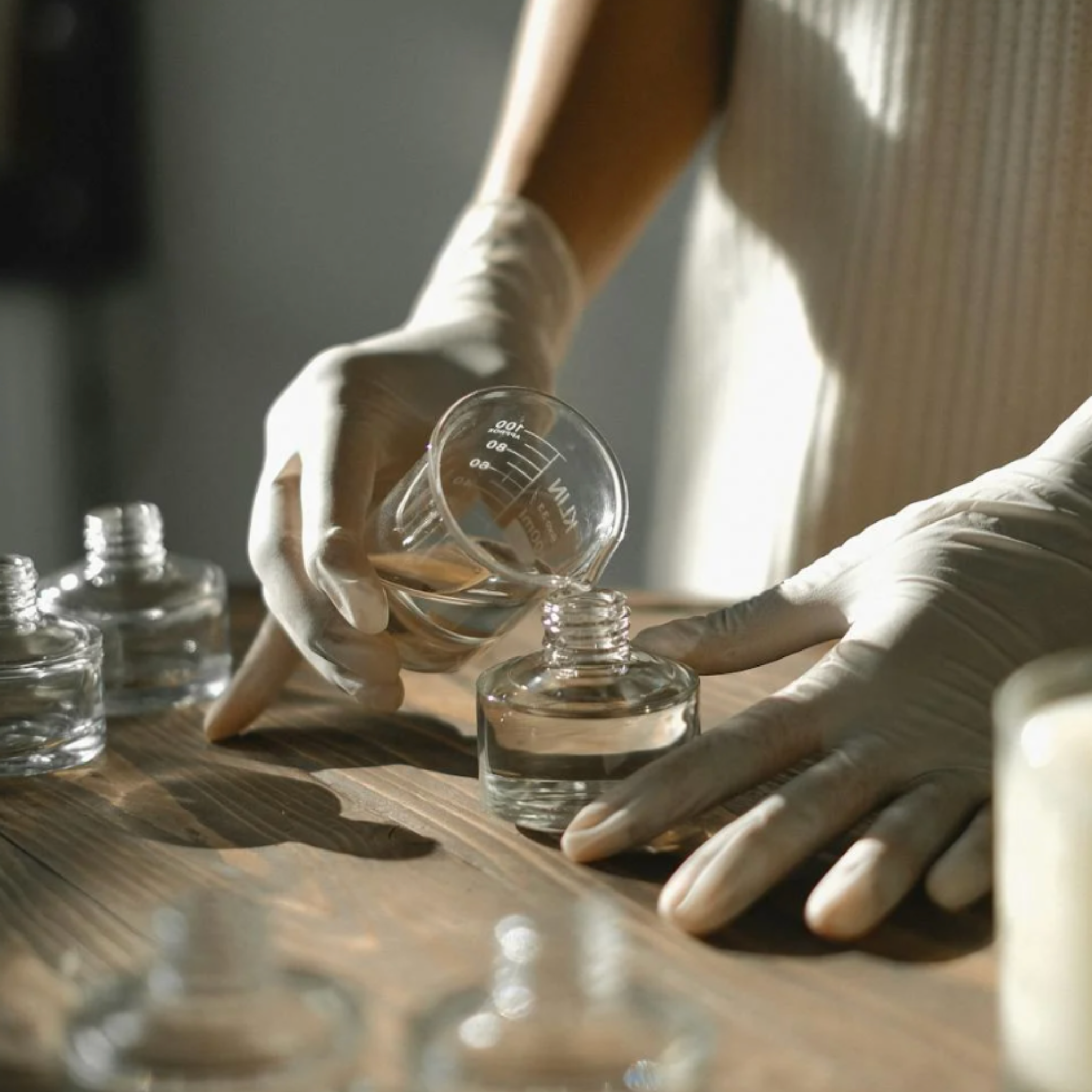 Person in white gloves pouring fragrance oil into a glass jar on a wooden surface