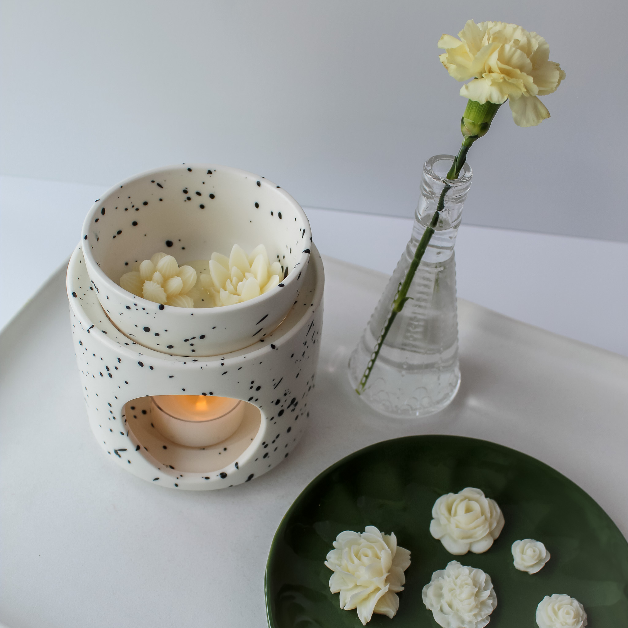 White wax warmer with black speckles beside a dark green dish holding five floral wax melts and a small vase with a single flower.